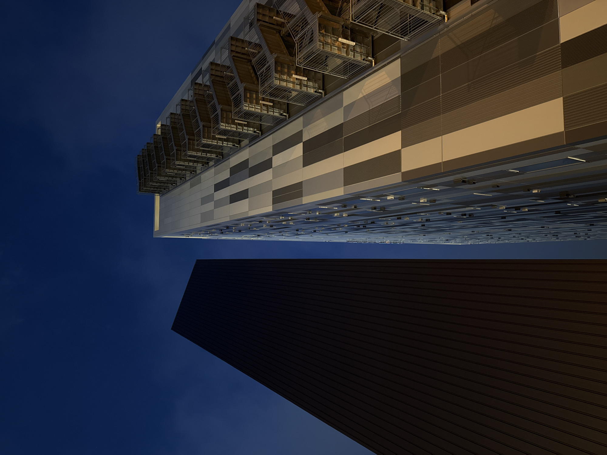 This media file features a low-angle perspective looking up at modern architecture against a dark blue, twilight sky. The primary subjects are two buildings. One building has a façade composed of horizontal panels in varying shades of grey and beige, with some sections featuring ribbed textures. Above this, a complex structure with exposed metal framework, possibly housing HVAC or support systems, is visible. The second building, appearing in the foreground and occupying the lower portion of the frame, is a large, dark, rectangular structure with a ribbed surface, angled upwards.
The time of day is clearly dusk or early evening, indicated by the deep blue of the sky and the artificial lighting not yet prominently illuminating the structures. There is no visible weather information. The architectural style suggests an urban or potentially industrial setting. No people or specific events are depicted. There is no visible text in the image. The overall impression is one of minimalist, imposing modern design.