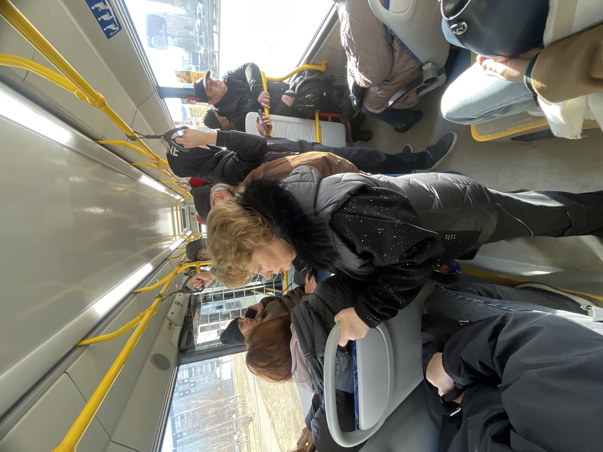 The image depicts the interior of a bus in Krasnoyarsk, Russia. The bus is crowded with passengers. There are yellow handrails along the top and sides of the bus. Passengers are standing and sitting. Some passengers are using their phones. The view from the windows shows a city landscape.