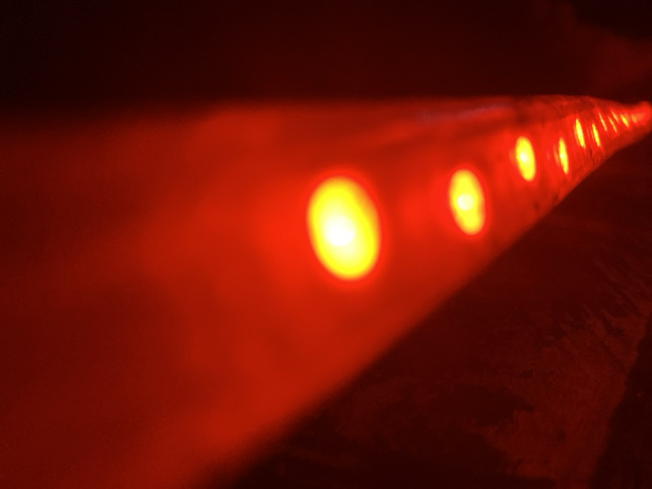 A low-angle, close-up image features a string of red lights with a yellow center. The lights recede into the distance, appearing smaller and closer together. The surface they are on appears dark. The color palette is primarily red and yellow, with the red hue dominating the overall scene. The lighting creates a dramatic effect.