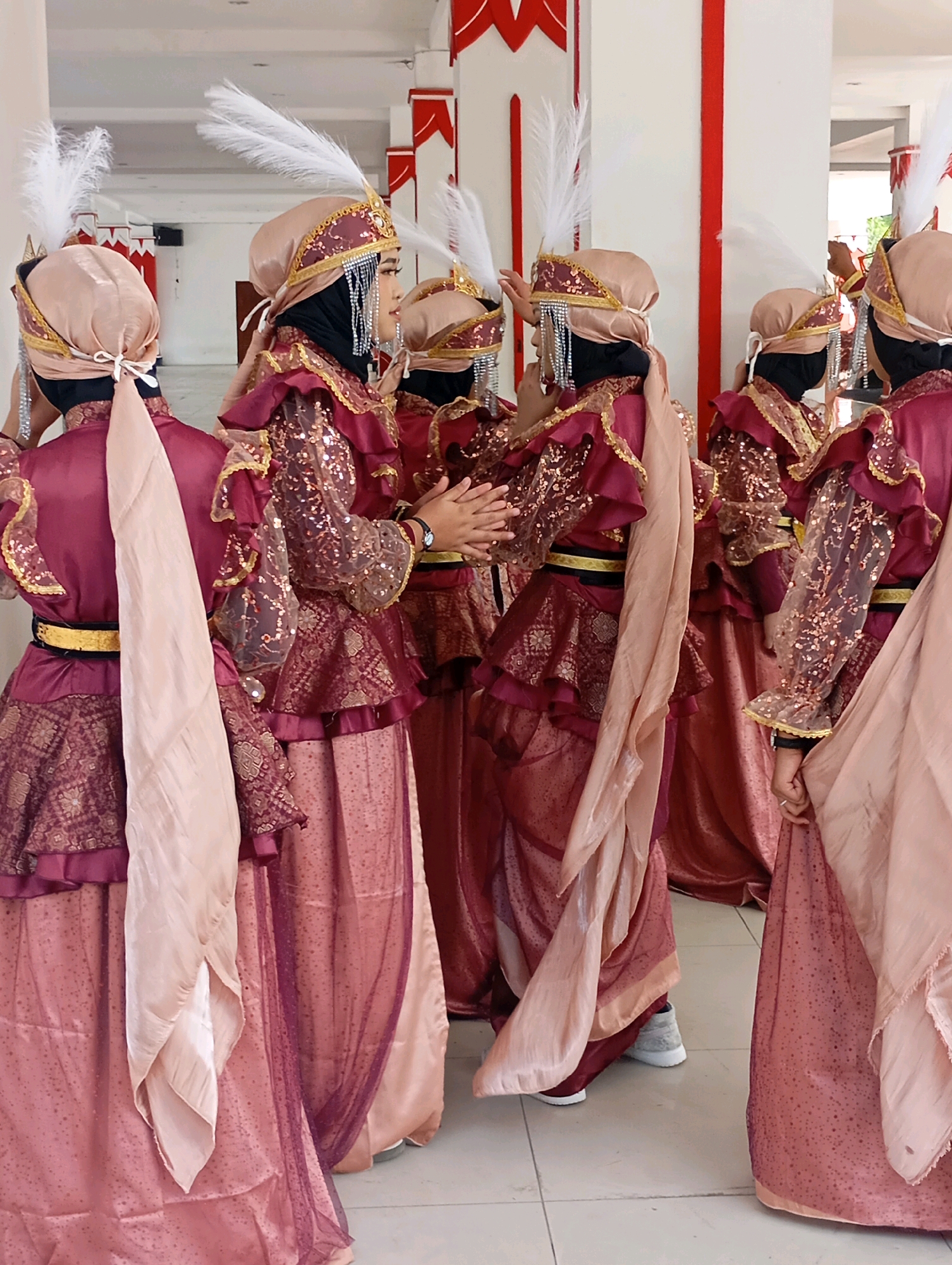 In Mungkid, Indonesia, a group of individuals in ornate, traditional attire stand in a hallway. The clothing consists of burgundy and rose gold garments with gold trim, intricate patterns, and sheer sequined sleeves. Each person wears a matching headscarf and a decorative headband adorned with white feathers and fringe. Some individuals have a pale pink sash draped over their shoulders. The background features white walls with red decorative elements, and the floor is tiled. One individual in the center is clapping their hands. The scene appears to be a cultural or performance-related gathering.