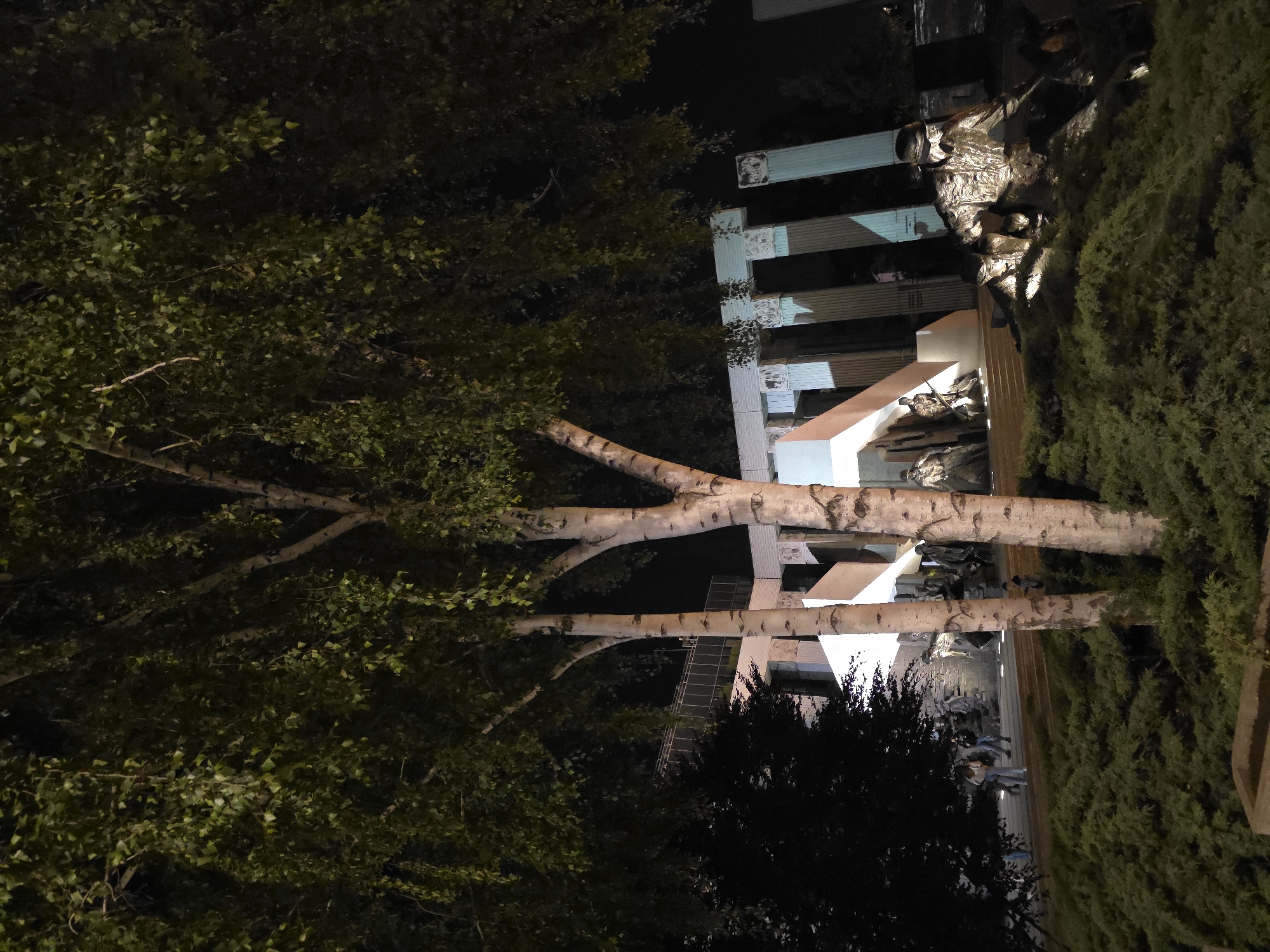 The image shows a nighttime view of the Warsaw Uprising Monument in Warsaw, Poland. The monument features a light-colored architectural structure with blue-green accents and is flanked by a copse of birch trees on one side and a lush green hedge on the other. Bronze sculptures depicting figures engaged in wartime activity are prominently displayed around the base of the monument. The scene is illuminated with artificial lighting.