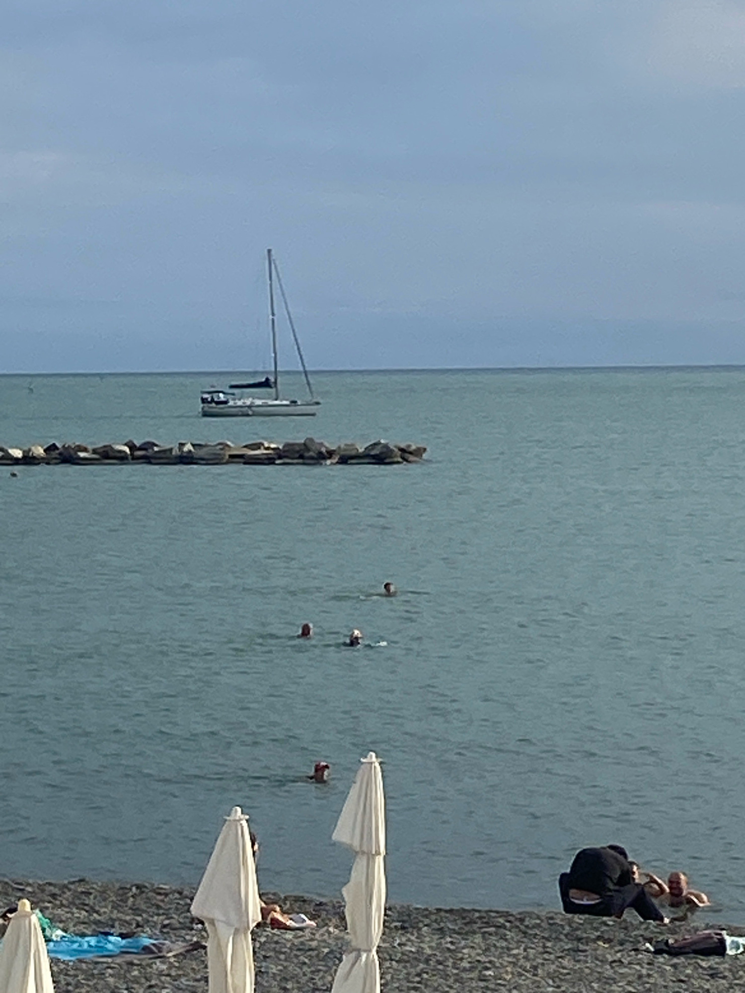 The image depicts a serene beach scene on an overcast day in Sochi, Russia. The sea is a calm, blue-green, with several people swimming in the distance. A white sailboat with a dark sail is anchored offshore, its mast reaching towards the cloudy sky. A line of large, grey rocks forms a breakwater in the middle ground. Closer to the shore, the beach is covered in dark pebbles. Several white beach umbrellas are set up, and a few people are relaxing on the beach, one in dark clothing is seated while another person is partially submerged in the water beside them. A blue towel and other beach accessories are visible on the pebbles near the umbrellas. The horizon line is clearly defined, separating the sea from the pale blue sky.