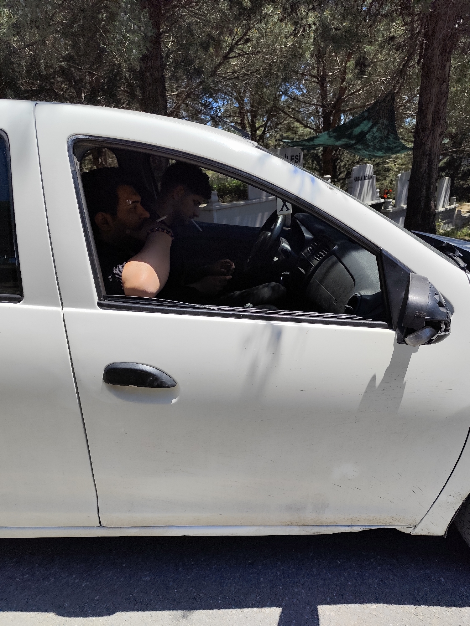 A medium shot captures the side of a white, four-door vehicle parked near a cemetery, possibly located in Sarıyer, Turkey, given the headstones with Turkish names visible through the car window and the style of the grave markers. Two male individuals are seated inside the vehicle. The driver has a cigarette in his mouth and is looking forward. The passenger, closer to the camera, has a beard and his right arm rests on the car door. The car shows visible signs of wear and damage, particularly to the side mirror, which is broken. The foreground shows a portion of the asphalt road, while the background consists of trees and headstones, with a green cloth or tarp draped over some of the grave markers. The time of day appears to be daylight hours.