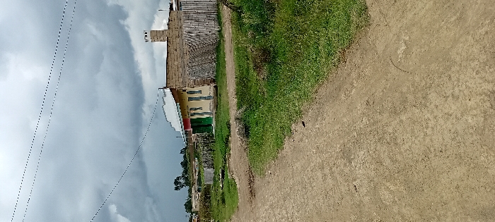 This image depicts a scene in the city of Heni Village, Kenya, under an overcast sky with dark grey clouds. The foreground consists of an unpaved, light-brown dirt road, flanked by patches of green grass and some dry vegetation on both sides. In the midground, a single-story building constructed from light-colored masonry, possibly painted a faded yellow-beige, stands prominent. Its front facade features a white, corrugated roof extension acting as an awning, two green double doors, and two vertically divided windows, all appearing closed. The building's right side is built with light-brown cinder blocks, and a brick chimney with a metal cap is visible on its roof. To the immediate right of this building, a fence or barrier composed of vertically arranged wooden planks or branches is present. Further in the background, to the left of the main building, additional single-story structures are partially visible amidst dense green foliage. Three parallel dark power lines are visible spanning across the upper portion of the frame against the cloudy sky. No individuals or vehicles are present in the scene.
