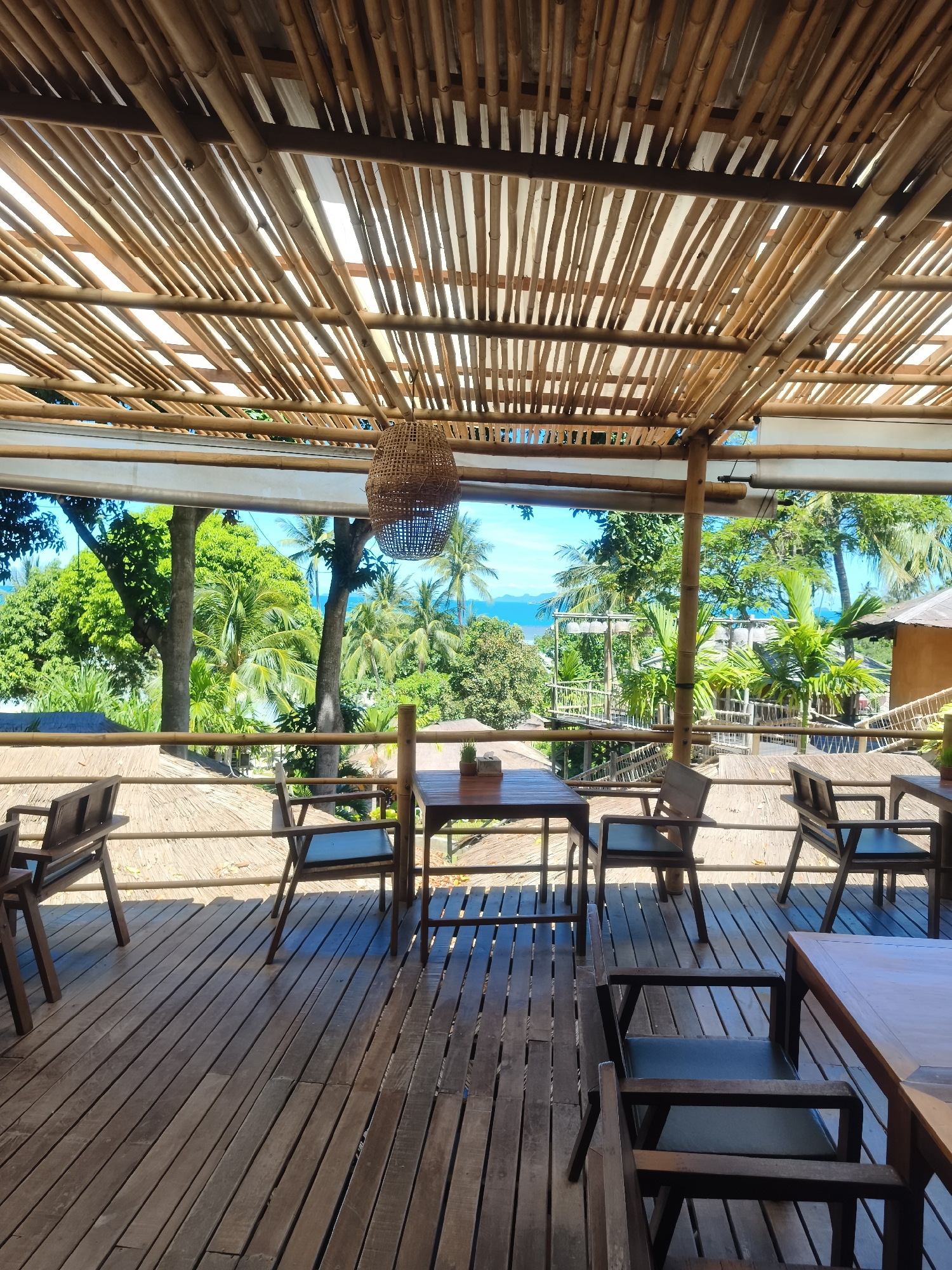 This is a vertical, medium shot of an outdoor dining area at Baan Thong Po, Thailand, suggesting a relaxed, tropical setting. The perspective is from within the covered area, looking outwards towards a vibrant green landscape and a bright blue ocean under a clear sky.
The foreground is dominated by a wooden deck with several tables and chairs arranged for dining. The furniture is simple and made of dark wood, with some chairs featuring dark cushioned seats. The tables are also wooden, with one in the center having a small potted plant on it. The deck itself is constructed from weathered wooden planks laid horizontally, with visible gaps between them.
Above, the ceiling is an intricate lattice of bamboo poles, allowing dappled sunlight to filter through. A woven basket-like light fixture hangs from the bamboo structure. A wooden railing separates the dining area from the view beyond.
Beyond the railing, lush tropical foliage is abundant, with tall palm trees and dense green trees filling the middle ground. In the distance, the turquoise ocean meets the bright blue sky, hinting at a coastal location. Some rustic structures, possibly part of the resort or other buildings, are visible amongst the trees, including a wooden walkway or bridge-like structure.
The scene appears peaceful and idyllic, likely during daylight hours given the bright sunlight and clear sky. There is no visible text. The overall impression is one of a serene, open-air dining experience in a tropical paradise.