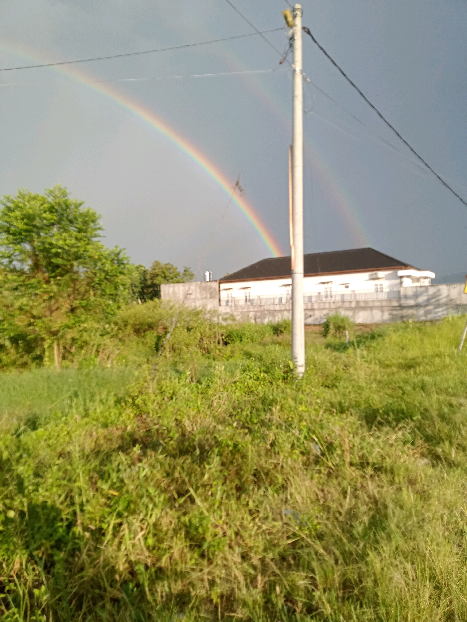 A wide shot shows a double rainbow arching across a gray sky over a suburban area in Hutagaol Peatalum, Indonesia. In the foreground, a patch of overgrown green grass and vegetation is visible. A utility pole stands to the right of center, with wires extending across the frame. In the middle ground, a white, single-story building with a dark roof is surrounded by a low concrete wall and fencing. The building appears to be a house or a small commercial structure. Lush green trees are also present on the left side of the image. The overall scene depicts a natural phenomenon occurring in a developed area.