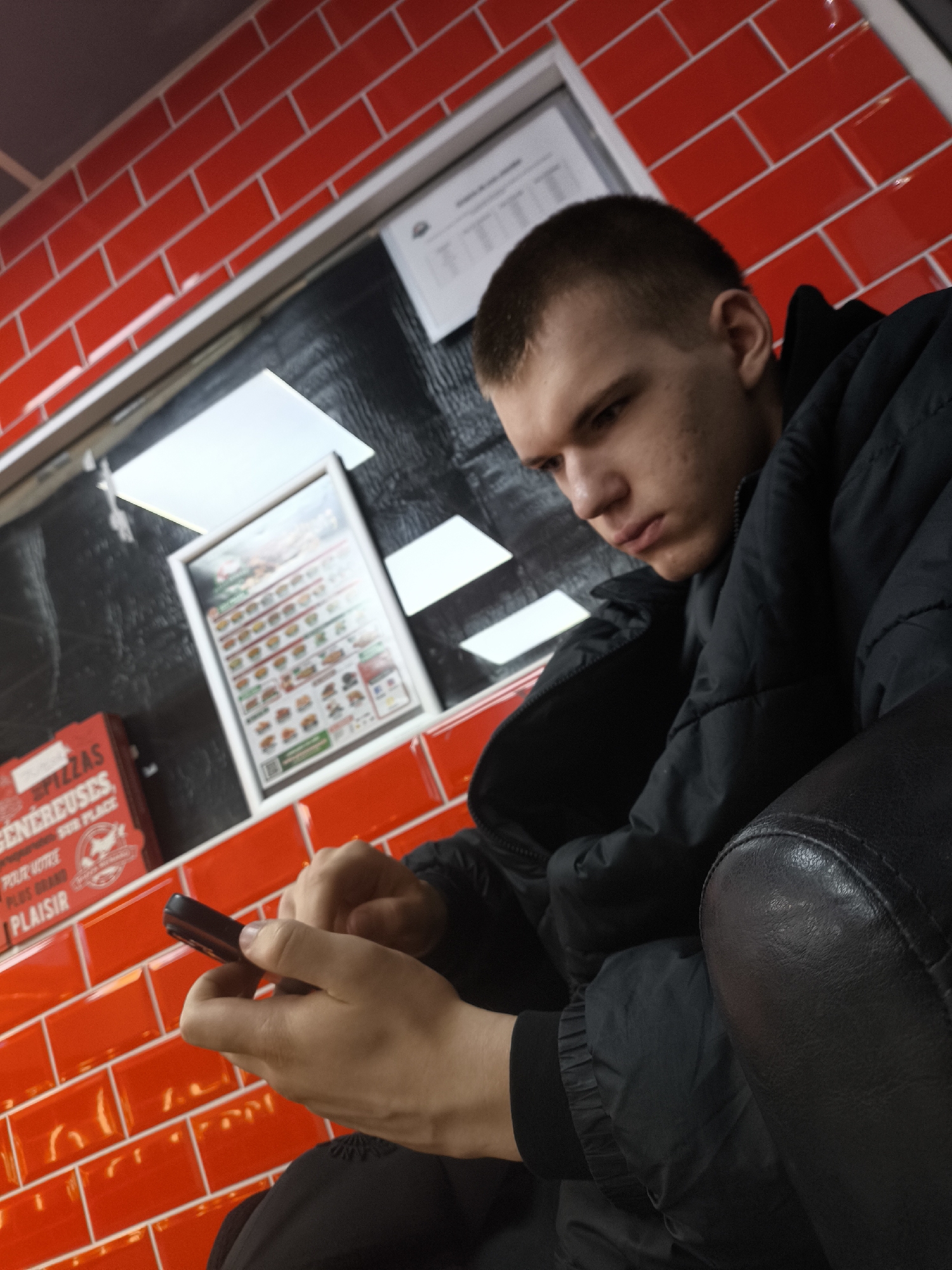 The image captures a young man seated, likely in a restaurant in Strasbourg, France, based on the "PIZZAS GENEREUSES" branding visible on the wall. The walls are covered in red brick tiles. The man is focused on his phone, which he holds in his hands. He is wearing a dark puffer jacket. A menu board is visible behind him. The image is taken from a low angle, suggesting the photographer is sitting or standing below him.