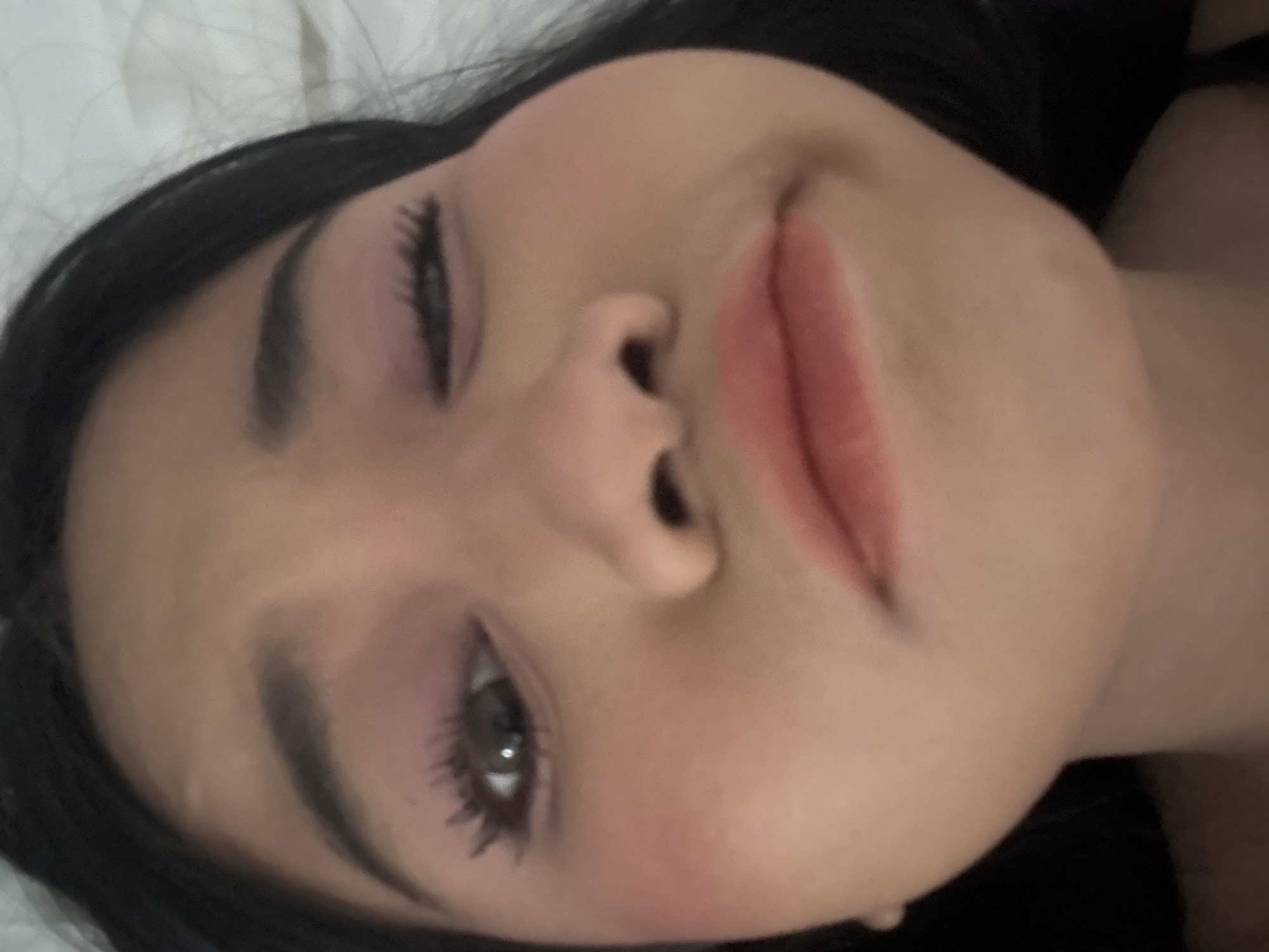 An extreme close-up portrait features a young individual, appearing female, captured making a playful or exaggerated facial expression. The subject's right eye (viewer's left) is wide open, gazing forward, while the left eye (viewer's right) is distinctly winking or nearly closed. The cheeks are noticeably puffed out, particularly on the subject's left side (viewer's right), and the lips are pursed and slightly downturned, angled towards the viewer's right. Dark hair frames the face. Visible makeup includes pink eyeshadow, mascara on prominent eyelashes, rosy blush on the cheekbones, and a reddish-brown lip tint. The individual has well-defined dark eyebrows. The background is uniformly light-colored and out of focus, suggesting an indoor setting with soft lighting, possibly bedding or a plain wall. The scene is presented as taking place in North Jakarta, Indonesia.