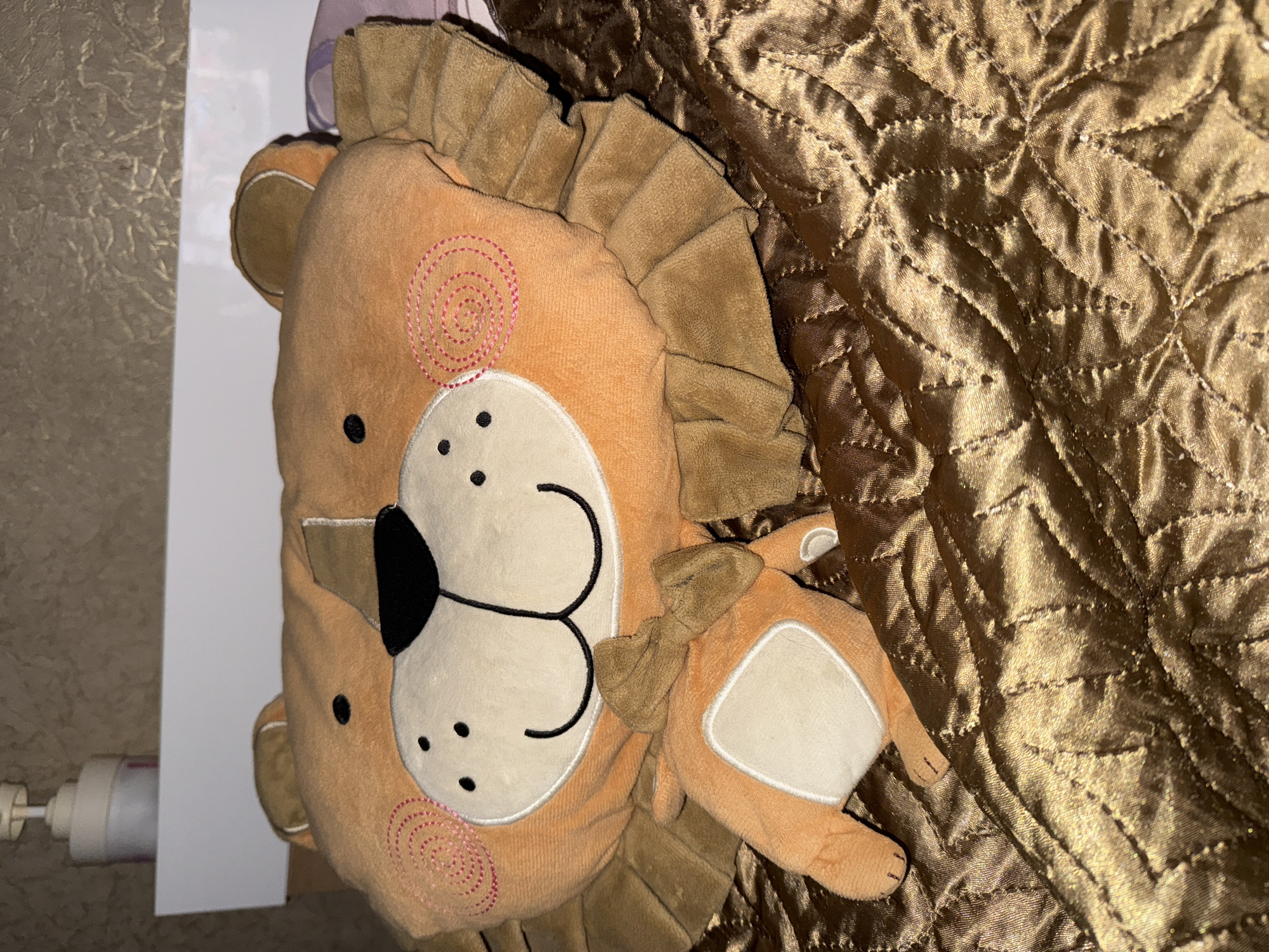 A stuffed toy lion is lying on a quilted, shiny brown blanket. The lion is orange with a brown mane and ears, and has a white face with black dots for eyes and nose, and a stitched smile. It is wearing a small brown bow tie. In the background, a white shelf holds a white pump dispenser. The wall behind the shelf has a textured, beige wallpaper. The image is taken from a slightly elevated angle, looking down at the toy. The scene appears to be indoors, likely in a bedroom. There is no indication of the location being in Novoshakhtinsk, Russia.