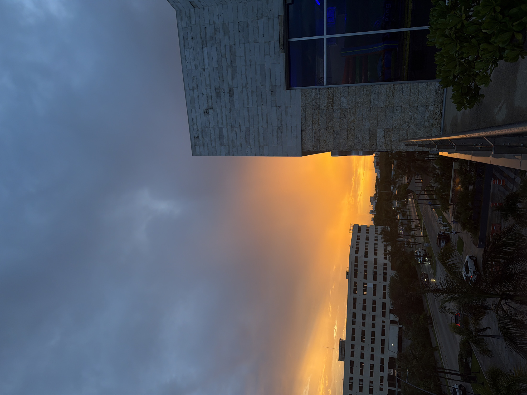 The image captures a sunset view in Cancún, Mexico, characterized by a dramatic sky with fiery orange and yellow hues blending into darker gray clouds. In the foreground, a modern building with stone cladding and large glass windows dominates the right side of the frame, with a balcony railing visible. To the left, a long, multi-story white building with numerous windows stretches across the mid-ground, with palm trees and streetlights lining the road in front of it. Vehicles, including cars and a van, are present on the road, suggesting an urban setting. The overall scene depicts a twilight atmosphere in a coastal city.