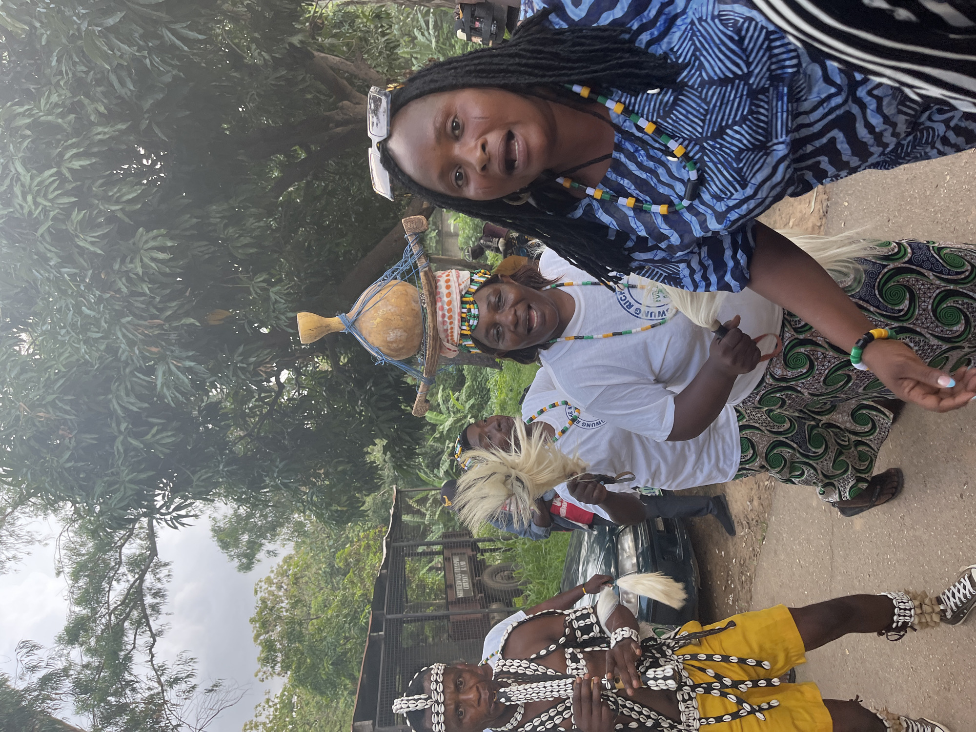 In Jos, Nigeria, a group of people are participating in a procession. The foreground is dominated by a woman with dark, braided hair, wearing a blue and black patterned dress and colorful beaded necklaces. She is looking directly at the camera with her mouth open as if speaking or singing. To her left, another woman wears a white t-shirt, a beaded headband, and a colorful skirt. She holds a white feathery object. Behind her, a man in traditional attire, adorned with shells and wearing yellow shorts, is holding a similar feathery object and playing a flute or pipe. He has shells around his head, neck, and wrists. In the background, more people are visible, some in white t-shirts and patterned skirts, and further back, a truck with a cage-like structure and an inscription that reads "SOUTHERN NIGERIA TRANSPORT COMPANY LIMITED." Trees and foliage form the backdrop, suggesting an outdoor, possibly rural or semi-rural setting within the city. The sky is overcast.