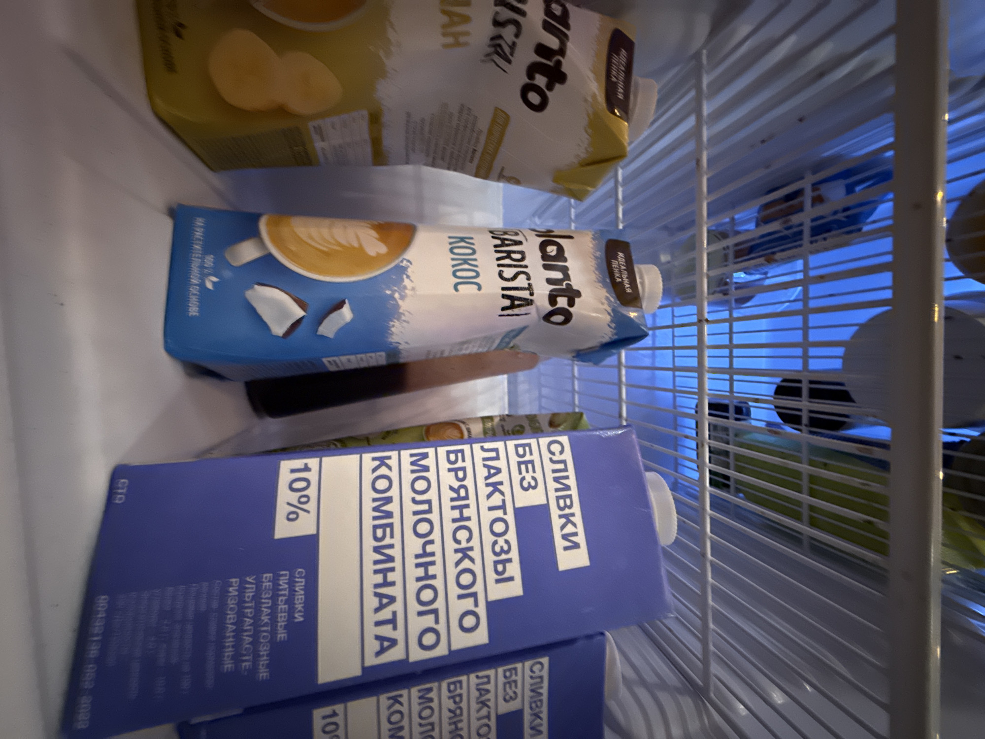 Inside a refrigerator, two cartons of milk-like beverages and another yellow carton are visible on a shelf. The carton on the left is yellow and appears to have text in a dark color, with an image of banana slices. The carton in the middle is white and blue, with "Planto BARISTA KOKOS" written on it. It also features an image of coffee with latte art and coconut pieces. To the right of this, a blue carton with white text reads "СЛИВКИ БЕЗ ЛАКТОЗЫ БРЯНСКОГО МОЛОЧНОГО КОМБИНАТА" (Cream without lactose from Bryansk Dairy Plant) and "10%". Another similar blue carton is partially visible to the far right. The shelves are made of white wire. The overall environment suggests a domestic refrigerator. The location is identified as Veshki, Russia.