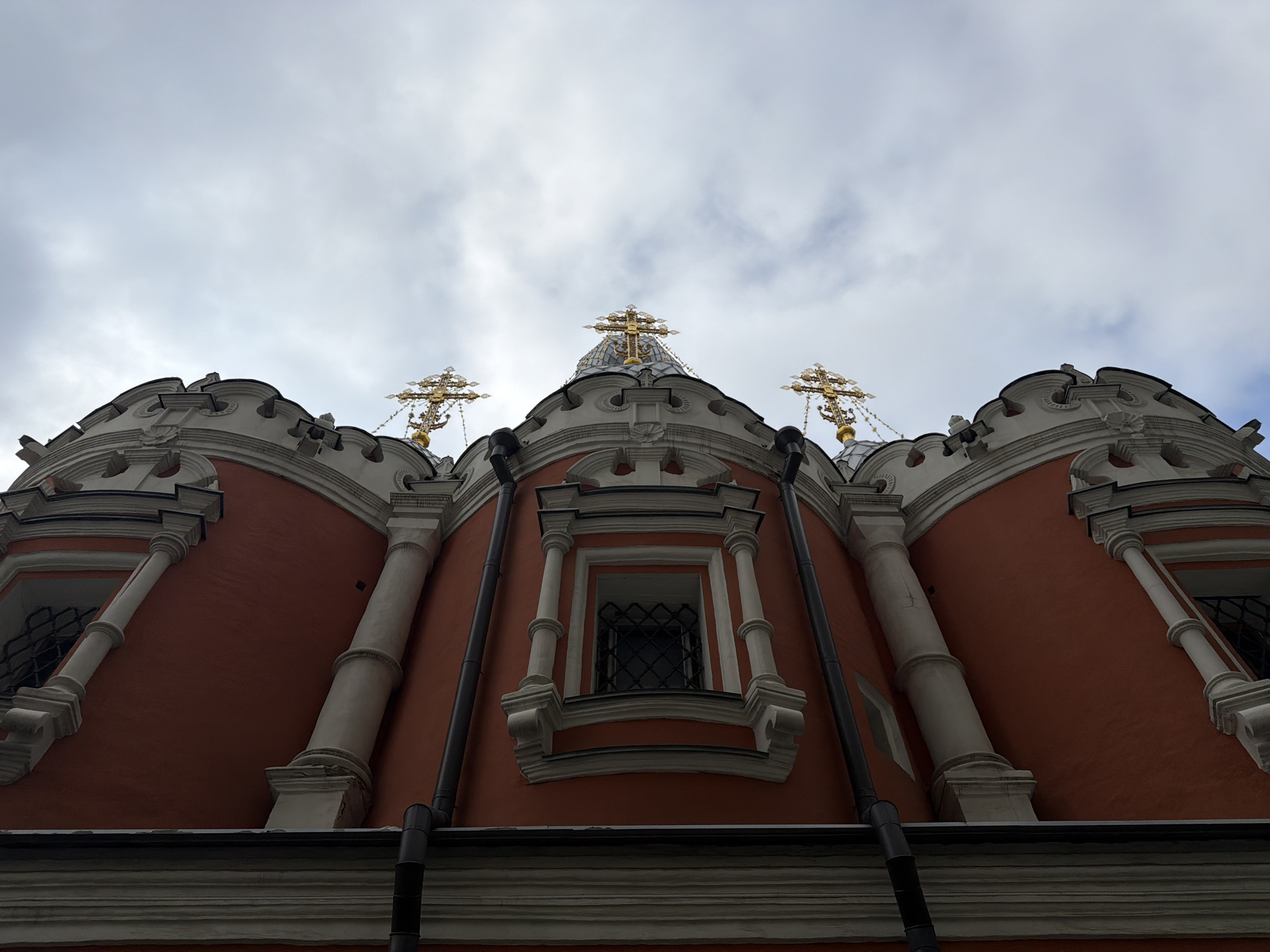 The image shows the upper portion of a red and white building under a cloudy sky. The building is likely a Russian Orthodox church, as indicated by the presence of three golden crosses atop small domes. The facade features decorative elements such as columns, window frames, and ornate trim. There is also a visible black downspout on the facade. This church is located in Moscow, Russia.