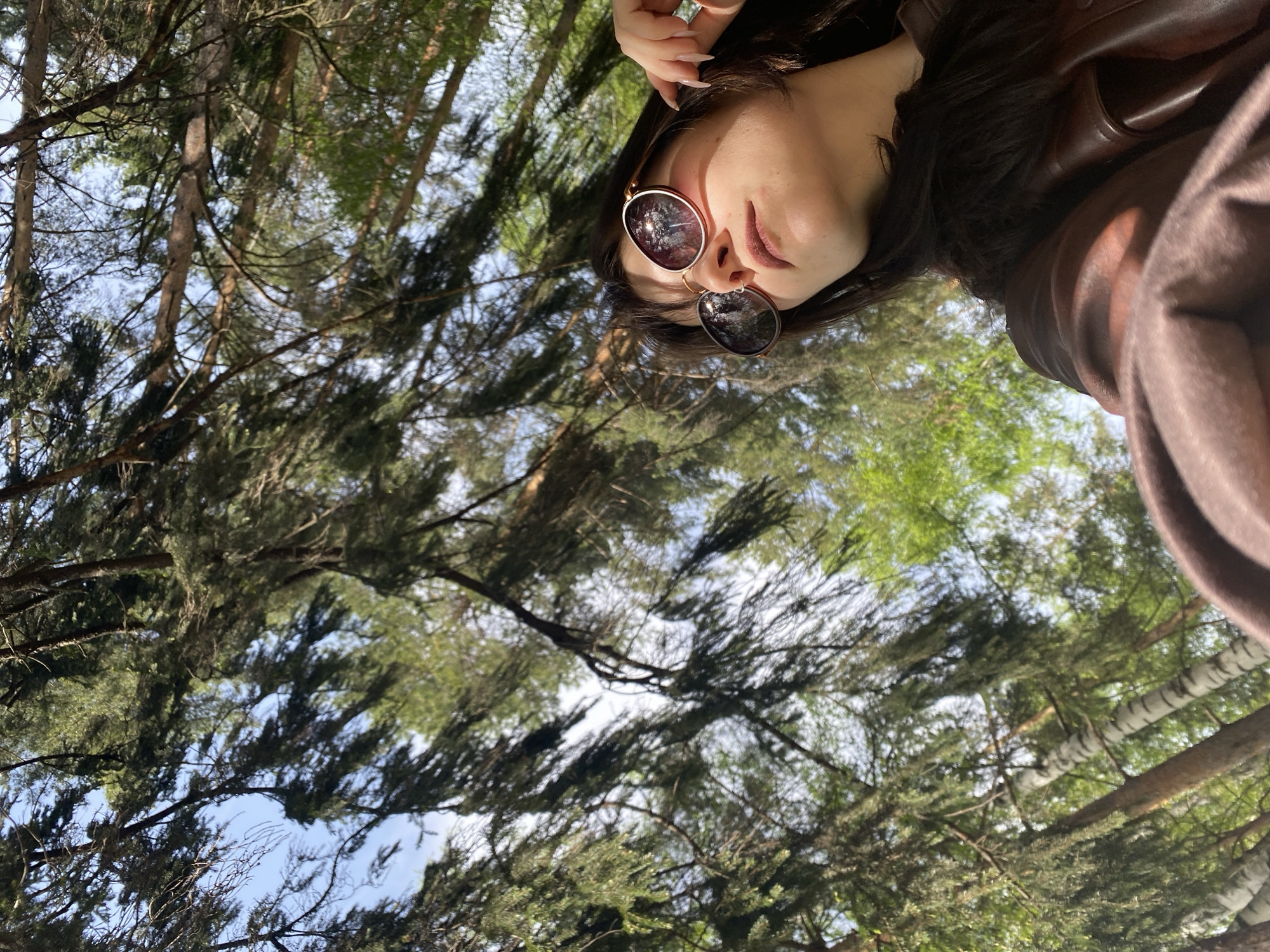 The image depicts a person lying on their back, looking up at the sky through trees. The person is wearing sunglasses and a brown jacket. They are also wearing a ring on their finger. The background is filled with trees and the sky is visible through the branches. The location is likely in a forest or park in Chaikovsky, Russia.