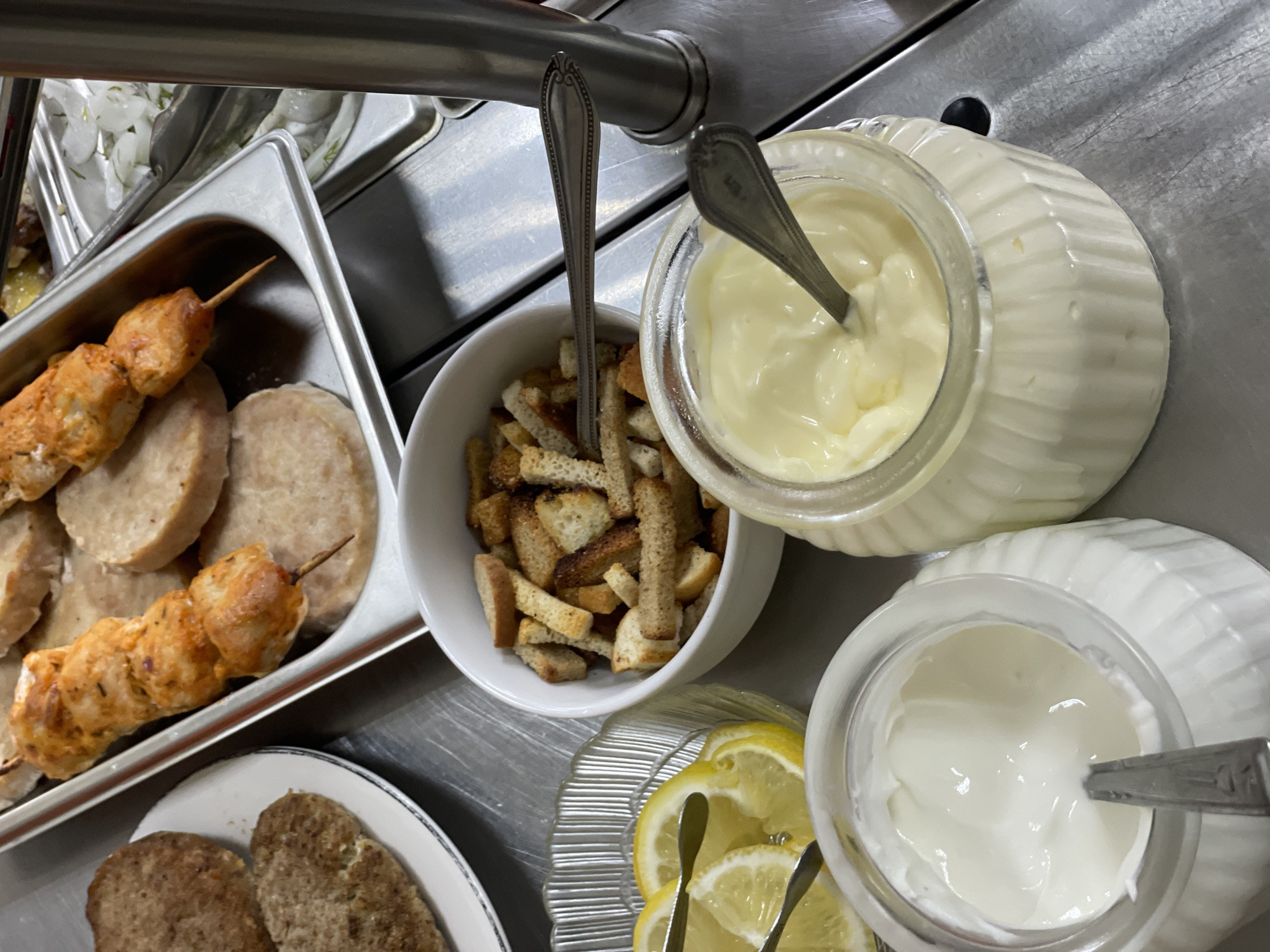 This media file shows a close-up, overhead view of a food service station, likely in a cafeteria or buffet setting. The scene is dominated by various food items arranged in metal and ceramic containers, set against a stainless steel countertop.
In the foreground, two glass jars filled with creamy white sauces are visible. One jar appears to contain mayonnaise, indicated by its slightly yellowish hue and the presence of a spoon. The other jar holds a whiter, thicker sauce, possibly sour cream or a similar condiment, also with a spoon resting inside. To the left of these jars, a white bowl is filled with croutons.
Further back, a rectangular stainless steel pan holds food on skewers. One skewer is clearly visible, featuring pieces of grilled chicken. Next to the skewers are several circular slices of a light-colored, processed meat, possibly ham or a similar luncheon meat.
In the bottom left corner, a white plate contains two oval-shaped, dark brown patties, which appear to be sausages or burgers. Adjacent to this plate is a decorative glass dish holding several thin slices of lemon, garnished with small metal tongs. In the background, a stainless steel trough holds what looks like a salad with onions and herbs.
The lighting suggests an indoor setting, likely illuminated by overhead lights, and the overall impression is one of food preparation or service. There are no people visible, and no discernible text. The focus is on the arrangement and presentation of the food items.