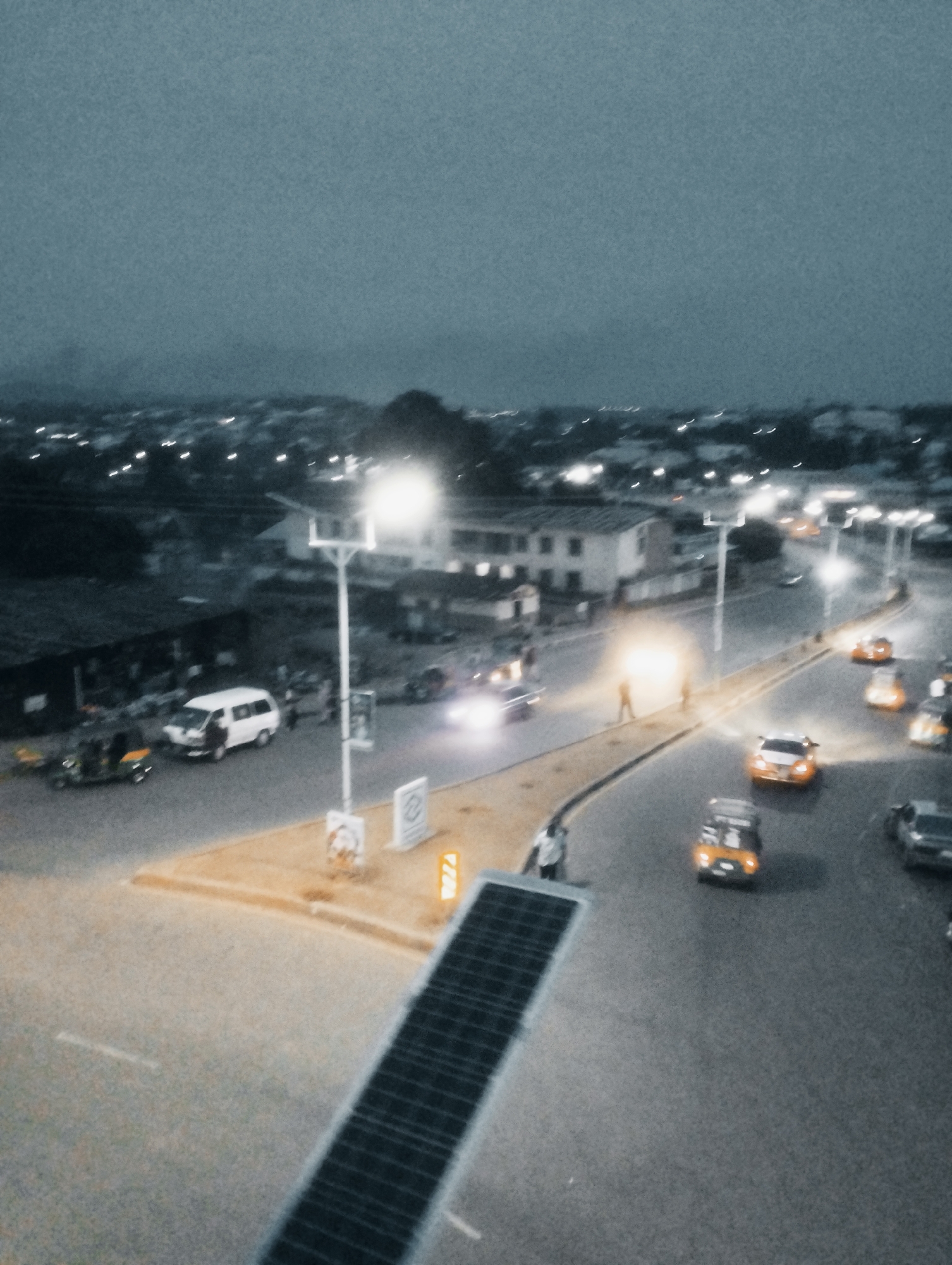 This media file depicts a street scene at dusk or night in Jos, Nigeria. The overall ambiance is dimly lit, with artificial lights from street lamps and vehicle headlights illuminating the road and surrounding buildings. The sky is dark and overcast, suggesting a cloudy evening.
The scene features a paved road with several vehicles, including cars and what appear to be minibusses or vans. Some vehicles are in motion, indicated by blurred headlights. There are also streetlights present, some of which are illuminated, and one particularly bright one near the center of the image.
On the side of the road, there are buildings, some appearing to be residential or commercial structures. Pedestrians are visible on the sidewalk and near the road, interacting with the environment. One notable object is a solar panel array positioned in the foreground, suggesting an interest in renewable energy.
The general atmosphere is one of a busy urban environment during a period of transition from day to night. The lighting conditions create a somewhat muted or moody visual, with some light sources creating glare. The presence of multiple vehicles and people suggests normal daily activity is ongoing. No specific event or gathering is clearly identifiable. There are no discernible words or text in the image, although some signs are present but unreadable due to low resolution and distance.