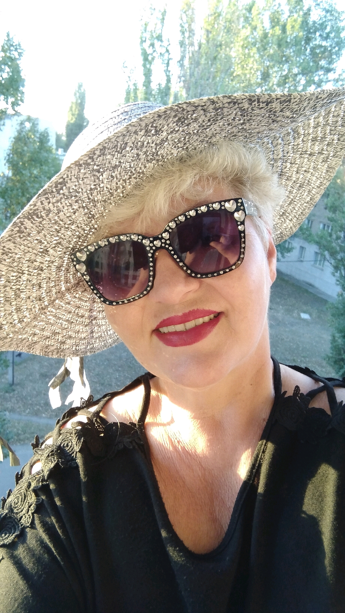 A woman with short, blonde hair is captured in a selfie outdoors, wearing a wide-brimmed, textured hat and oversized sunglasses adorned with rhinestones. Her lips are painted a bright pink. She is dressed in a black top with lace detailing on the shoulders and straps. The background suggests an urban environment with trees and buildings visible. The lighting indicates it is daytime, possibly late afternoon, given the strong shadows. The image appears to be taken from a slightly elevated position, possibly a balcony or window. The city of Horishni Plavni, Ukraine, is a plausible location given the urban setting and vegetation.