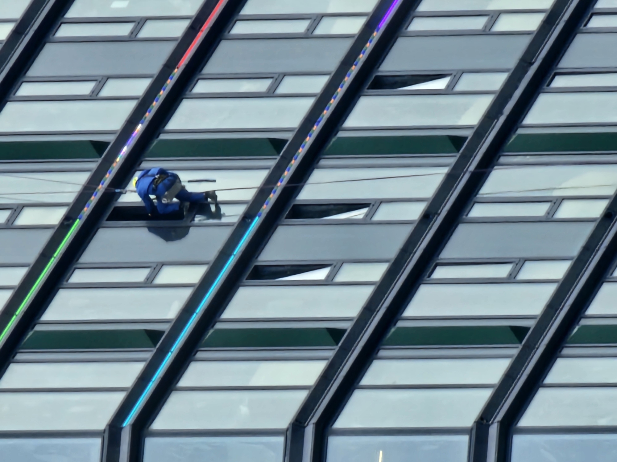 A person in a blue jumpsuit and yellow hard hat is rappelling down the side of a modern building. The building has a facade of reflective glass windows and is adorned with colored LED lights along horizontal ledges. The person appears to be cleaning or inspecting the windows. The image is taken from a low angle, looking up at the building. The sky is overcast. This image was taken in Moscow, Russia.