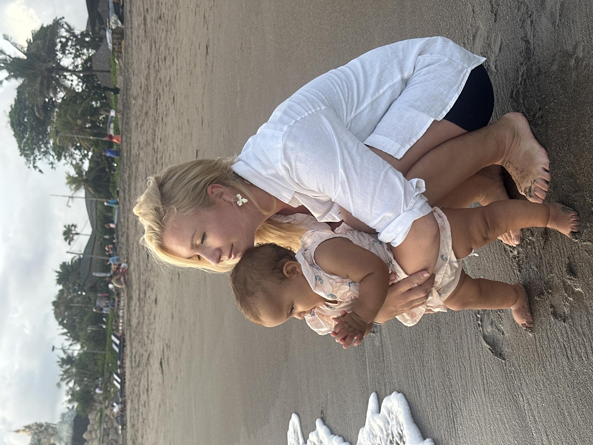 A woman and a baby are on a beach in North Kuta, Indonesia. The woman, with blonde hair and fair skin, is kneeling, wearing a white shirt and black shorts. She is holding the baby, who has brown hair and skin, near the edge of the water. The baby is wearing a light pink romper with an elephant print. The beach is sandy and wet, with gentle waves coming to shore. In the background, there are palm trees and buildings along the coastline.