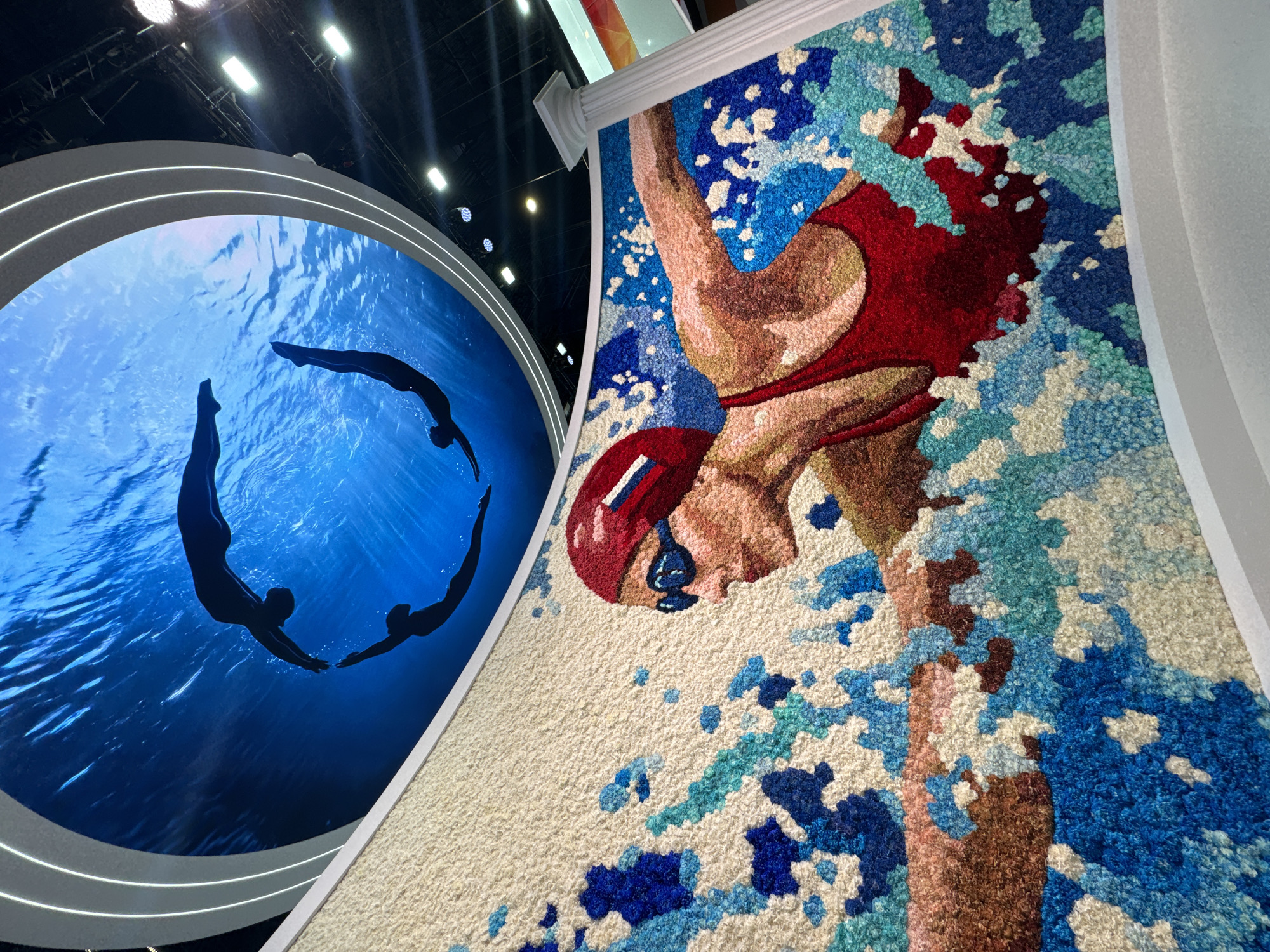This media file captures a vibrant indoor installation in Shushary, Russia, featuring two large, distinct artworks centered around aquatic themes.
On the left, a large, circular display presents an immersive underwater scene. Four silhouetted human figures, possibly synchronized swimmers or divers, are depicted against a bright blue background, illuminated by natural light patterns filtering through the water's surface. Two figures are positioned above, diving downwards, while the other two are below, swimming horizontally in a flowing, circular formation. The display is framed by illuminated white rings, contributing to a modern aesthetic.
On the right, a large, textured artwork, resembling a rug or tapestry, portrays a swimmer in motion. The artwork is composed of countless small tufts of yarn or fabric, creating a tactile, pixelated effect. It depicts the upper body of a swimmer with raised arms, wearing a red swim cap, blue goggles, and a red swimsuit, set against an abstract background of various shades of blue and white, evocative of water splashes and bubbles. The swimmer's skin tone is rendered in shades of brown and tan. This vertical artwork is bordered by a white, classical architectural molding.
The scene appears to be an exhibition or public art space, characterized by artificial overhead lighting and clean architectural lines. Both displays emphasize dynamic movement and artistic interpretation of aquatic activities. There is no visible text in the image.