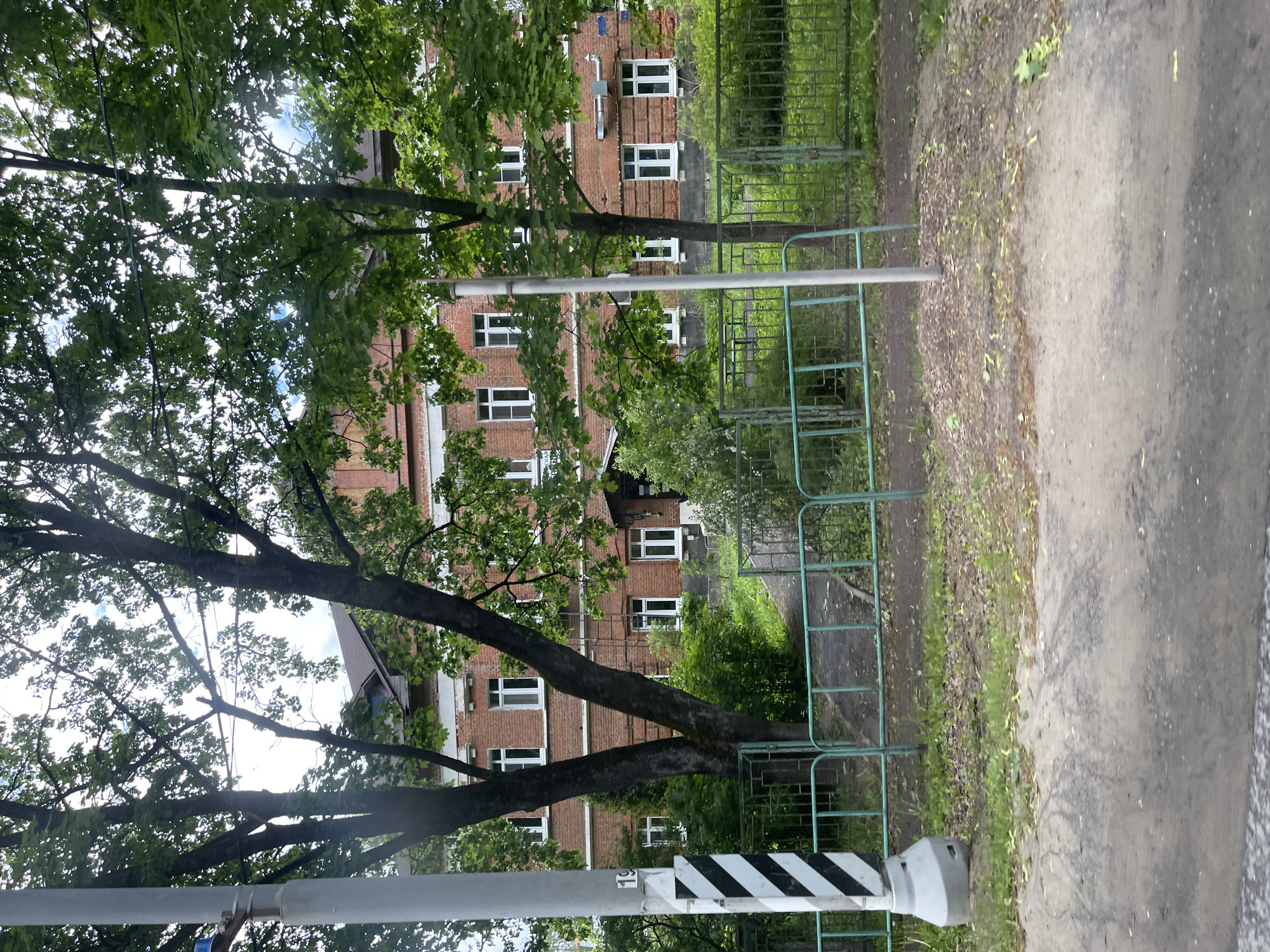 The image shows a street scene in Semyonovskoye, Russia. A red brick building with multiple white-framed windows is visible behind trees with green foliage. A green metal fence runs along the right side of the frame, bordering a paved pathway. A gray post with black and white striped markings stands in the foreground. The scene appears to be outdoors during daylight. No people are visible.