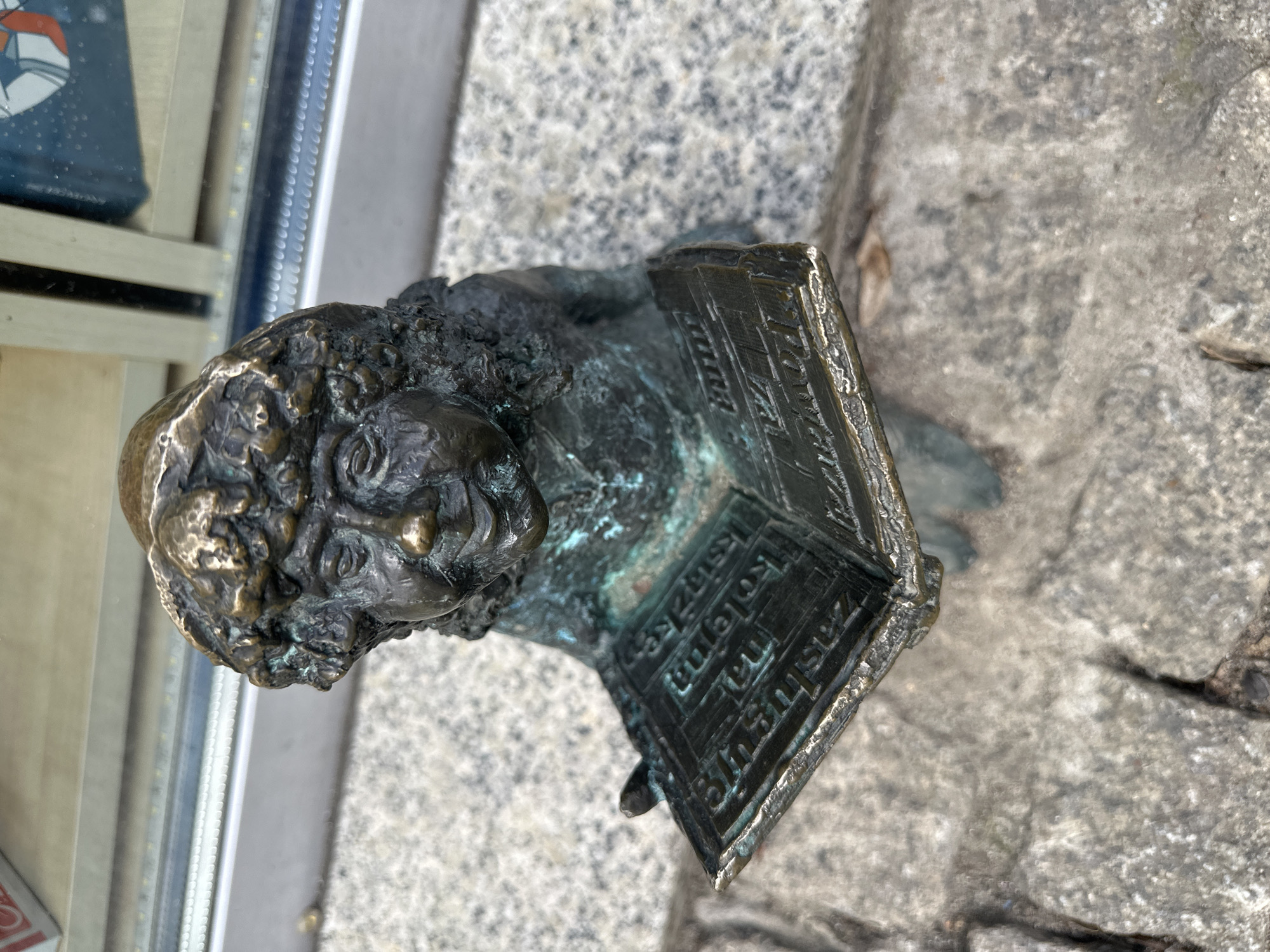 A high-angle, medium shot shows a bronze dwarf statue in Wrocław, Poland. The dwarf is sitting and reading a book. The book has Polish text on it. The statue is on a stone surface. A glass window is visible in the background to the left.