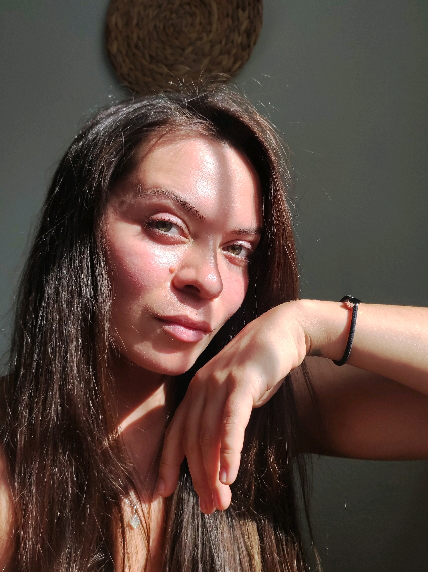 A young woman with long, dark brown hair is captured in a close-up, head-and-shoulders portrait. She is looking towards the viewer with a neutral expression. The lighting suggests she is indoors, with strong directional light creating shadows across her face and hair. Her left arm is bent and resting on her cheek, with a thin black bracelet visible on her wrist. A small, light-colored pendant necklace is also visible. The background is a plain, dark gray wall with a woven circular object hanging near the top. The image does not contain any information that would allow for the identification of its location in Chaikovsky, Russia.