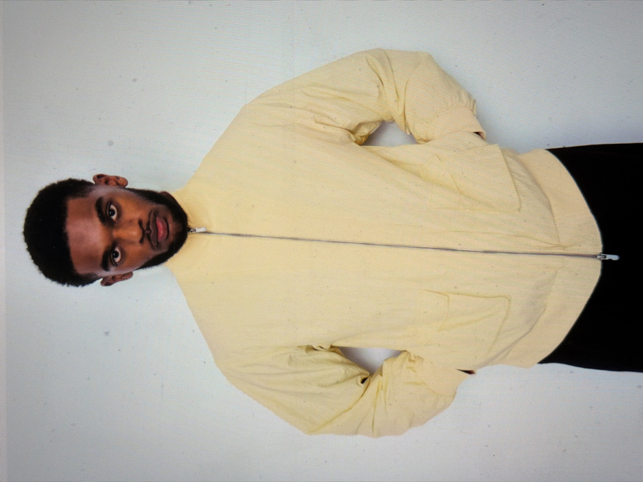 A single male subject, appearing to be a young adult of Black ethnicity, is depicted standing centrally against a plain, light-colored wall. He has short, dark, curly hair with faded sides and a neatly trimmed dark beard and mustache. The subject is wearing a light yellow, long-sleeved jacket featuring a high collar, a full-length silver zipper closure, and two lower front flap pockets. His hands are placed into the jacket's side pockets. Dark trousers are visible below the jacket's hem. The subject maintains a direct gaze towards the viewer with a neutral facial expression. The background offers no distinguishing features, environmental context, or architectural elements to indicate a specific location within the city of London, United Kingdom.