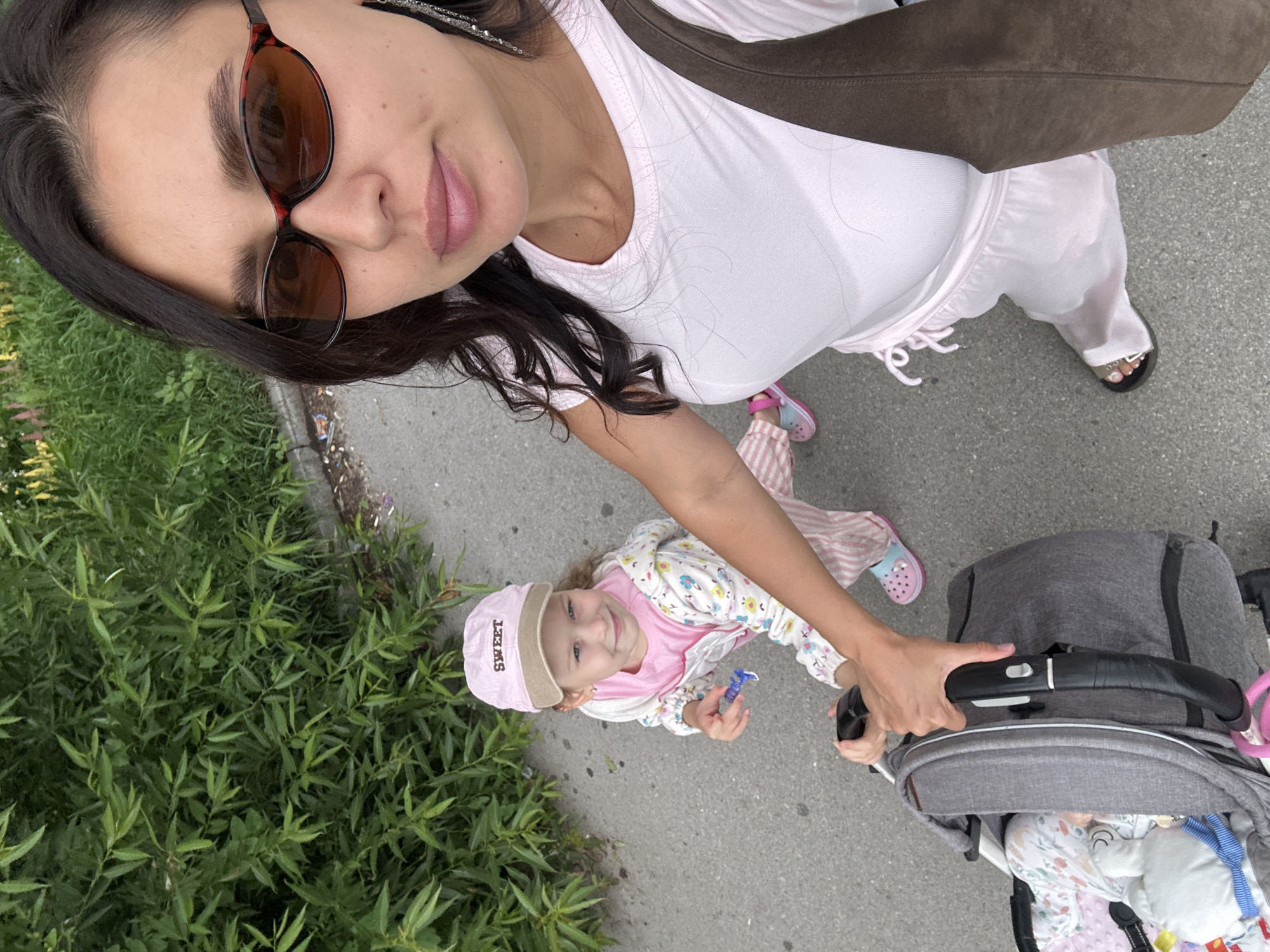 In the city of Perm, Russia, a woman is taking a selfie with her young daughter and a stroller. The woman, wearing sunglasses and a pink top, is in the foreground, with her daughter, who is wearing a pink cap, and a stroller visible behind her. The background consists of lush green bushes and a paved walkway. The daughter is reaching for the stroller handle, and the woman's hand is also on the handle. The scene appears to be an outdoor setting, possibly a park or a residential area.
