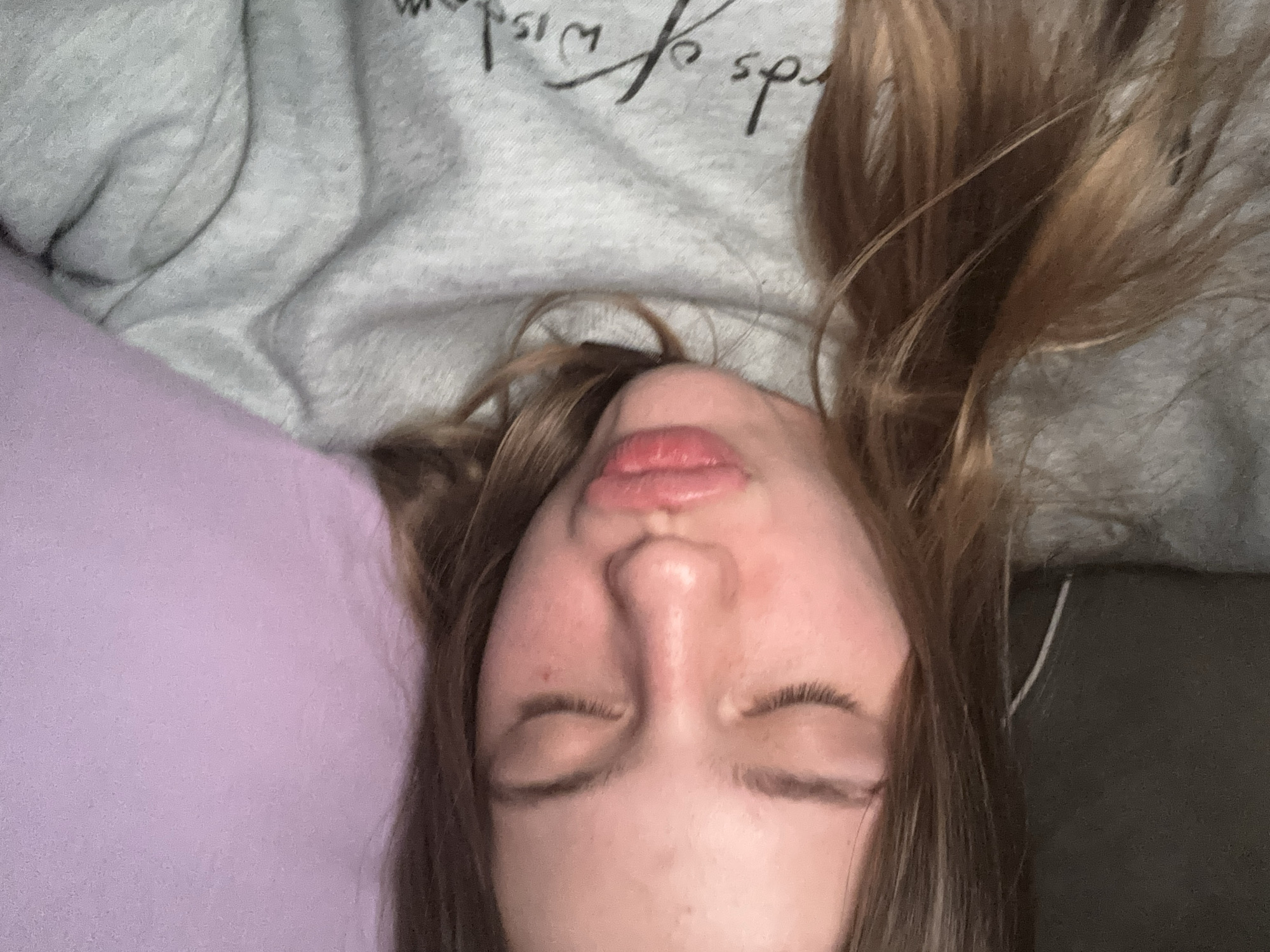A young woman with long, light brown hair and closed eyes is captured in a close-up selfie. Her lips are pursed, and her skin appears flushed. She is wearing a grey sweatshirt with the words "Words of Wisdom" partially visible on the front. The background consists of a textured purple wall. The image appears to be taken indoors, likely in a residential setting, and there is no indication of the city of Ladozhskoe Ozero, Russia.