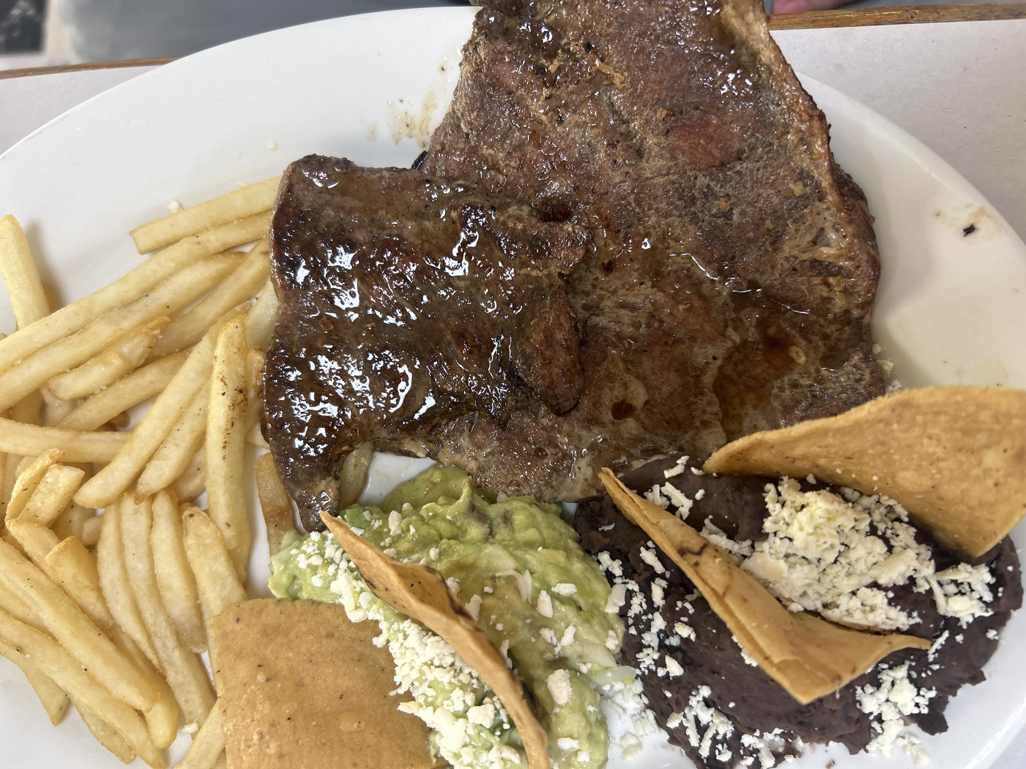 A plate of food sits on a white surface. The food consists of a steak, a pile of french fries, a portion of guacamole topped with cheese, a portion of refried beans topped with cheese, and tortilla chips. The steak appears to be coated in some sort of sauce. The location is presumed to be Puebla, Mexico. No people are visible.