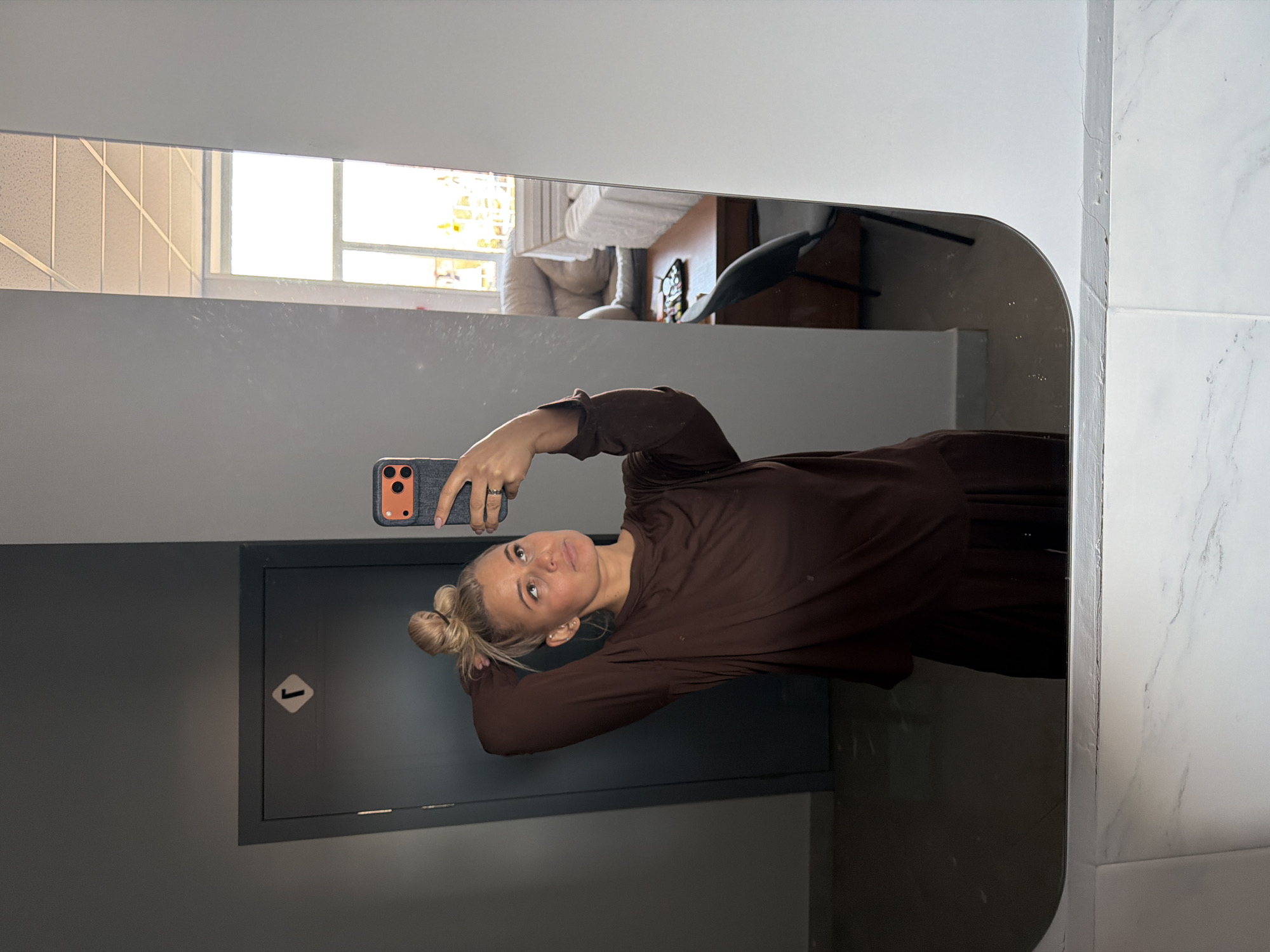 The media file displays a mirror selfie, rotated 90 degrees clockwise from its natural orientation. The primary subject is a young woman with blonde hair, styled in two buns. She wears a long-sleeved, loose-fitting dark brown top or dress and a ring on her left ring finger. She holds a smartphone with a grey textured case and an orange accent around the camera module, directing it towards the mirror for the photo. Her expression is neutral as she looks towards her reflection.
Reflected in the mirror is a brightly lit indoor space, suggesting it is daytime. A large window with multiple panes reveals strong daylight. Furniture, including a light-colored sofa or armchair and a dark chair, along with a grid-patterned ceiling, are visible in the background reflection.
In the immediate surroundings where the photo was taken, a plain grey wall is visible, alongside a dark grey panel or door. This door features a white square sticker with a black upward-pointing arrow. On the far right, a wall covered with white marble-patterned tiles borders the mirror. The area directly in front of the mirror, representing the floor, appears dark and reflective.
The clean, modern aesthetic with the architectural elements, such as the styled hair buns, and the door sign, suggest an indoor public or commercial environment like an office building, shopping center, or a hotel in Sarapul, Russia. The woman is actively engaged in taking a mirror selfie.