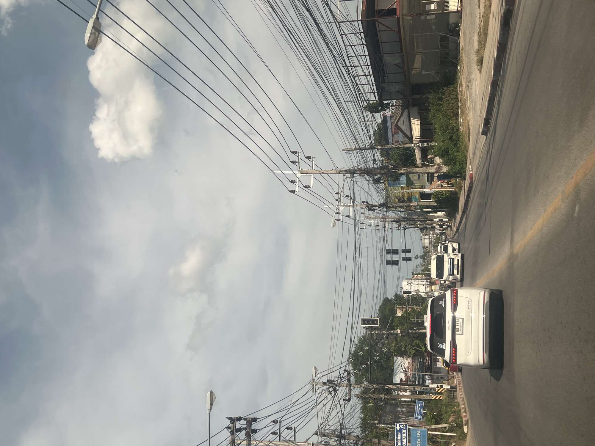 The image captures a view from inside a vehicle traveling on a road in the Ko Samui District of Thailand. The sky is overcast with scattered clouds. The road is paved and has yellow and white lane markings. On the left side of the road, there are signs, including one that reads "CONDUMINUM" and another for "GITA SPA". Utility poles with a dense network of electrical wires are prevalent on both sides of the street. Trees and various buildings line the roadside, some appearing to be under construction or renovation with scaffolding. Several vehicles are visible on the road, including a white SUV in the foreground with a license plate displaying Thai characters, and white vans and other cars further ahead. Traffic lights are present at an intersection. The overall scene depicts a typical urban or semi-urban environment in Thailand, characterized by heavy utility infrastructure and a mix of commercial and residential buildings.