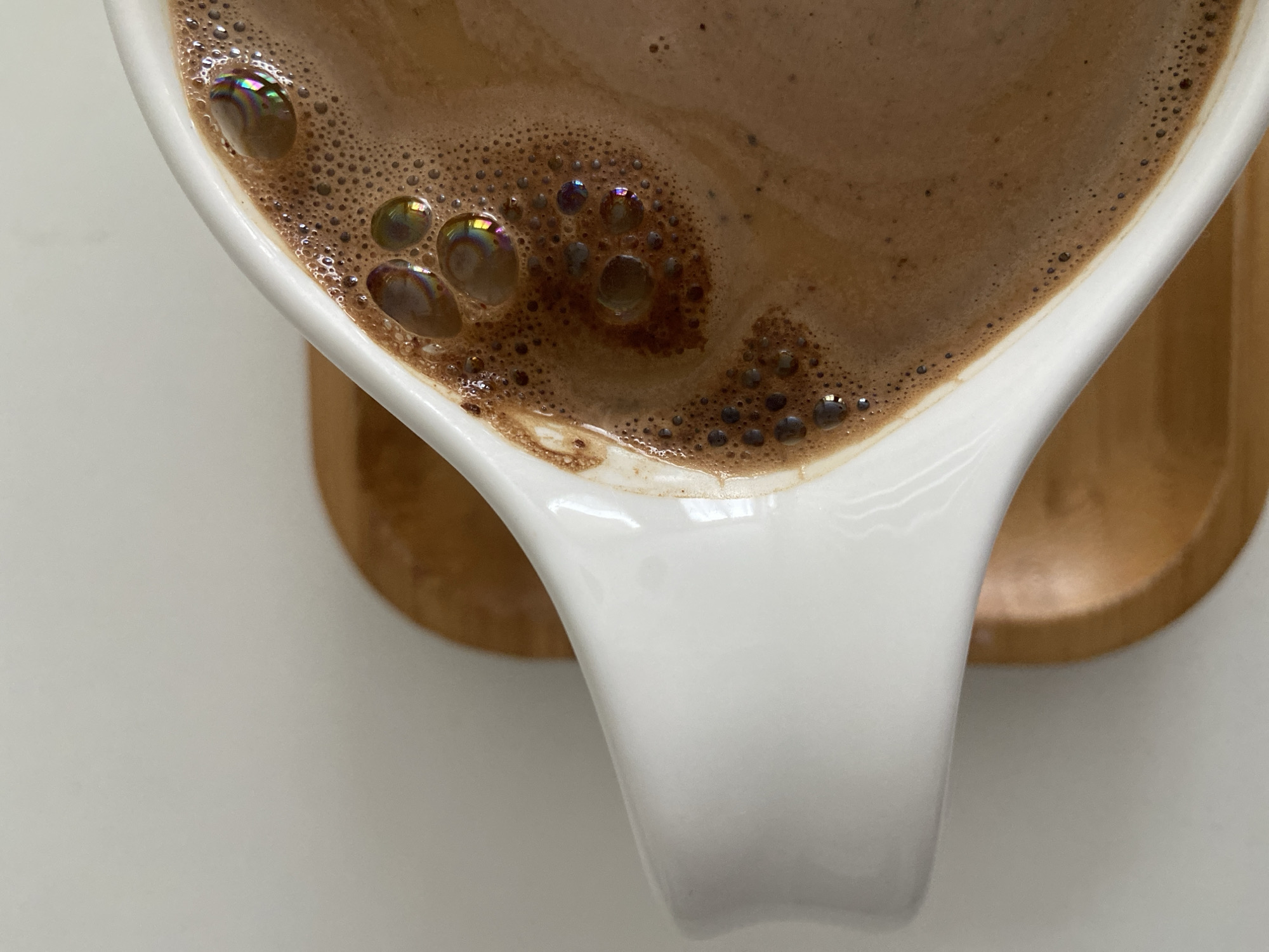 An overhead shot focuses on a cup of coffee or hot chocolate. The beverage is brownish, with numerous small bubbles and larger iridescent bubbles near the white ceramic cup's edge. The cup sits on a light brown wooden coaster, which is placed on a white surface. The scene lacks any indication it is in Budva, Montenegro, or features any people or specific actions.