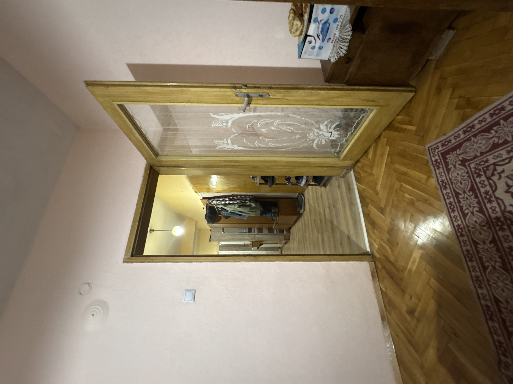 The image depicts a doorway opening into what appears to be a closet or small room. The doorway is wooden with a glass or translucent panel featuring a floral or decorative pattern. The room seen through the doorway contains shelves or storage units with clothes and other items visible. The walls are painted in a light pink color. The floor in the foreground is wooden parquet. A rug with a red and white pattern is partially visible on the right side of the image. The location is in an indoor setting, but the city of Radomir, Bulgaria cannot be determined from the image.