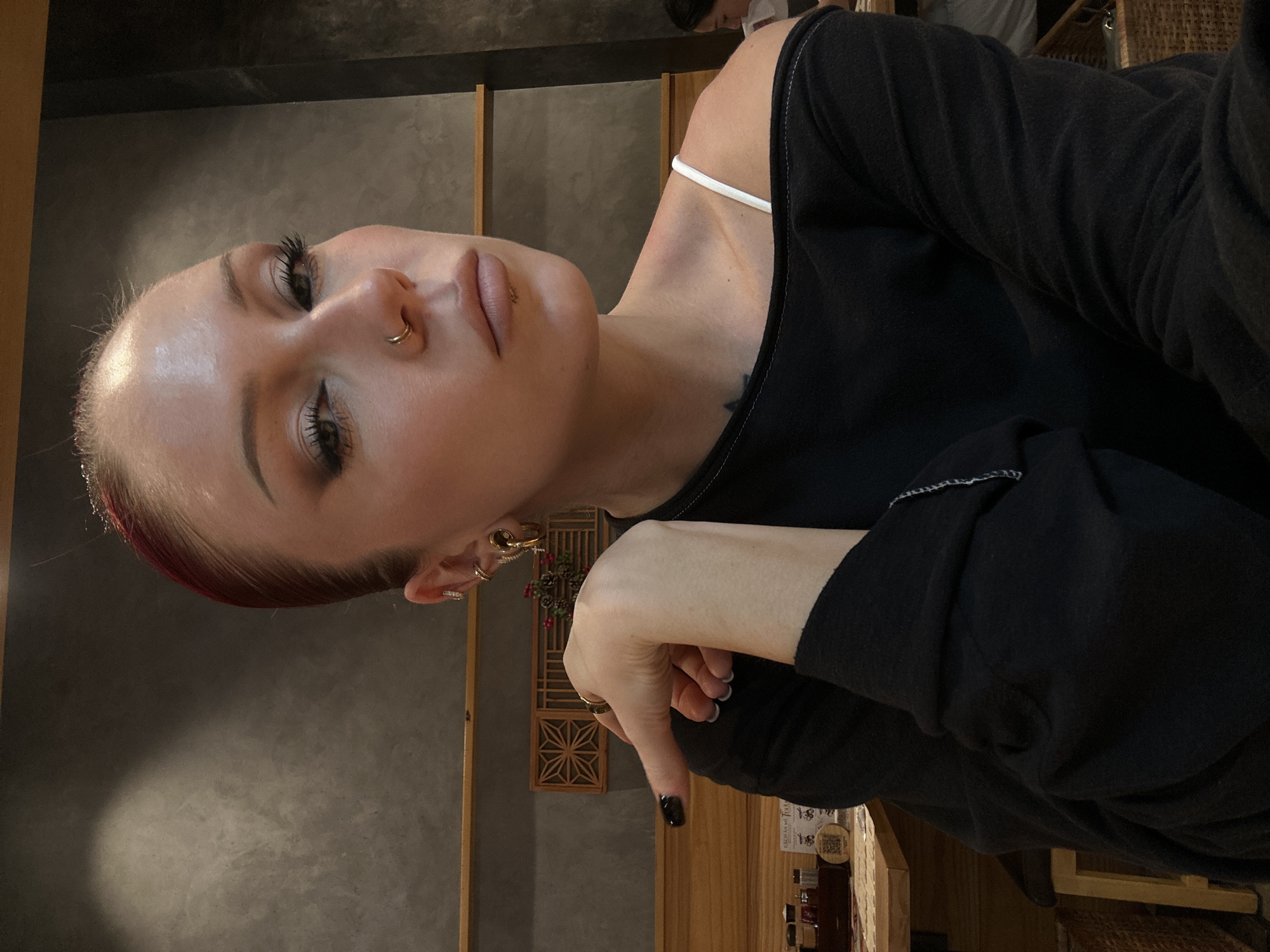 A young Caucasian woman with dyed red hair pulled back, wearing a black off-the-shoulder top and a white spaghetti strap shirt underneath, poses for a portrait in a restaurant in Da Nang, Vietnam. She has makeup on, including dark eyeliner and mascara, and a nose ring. Her left arm is bent, with her hand resting on her cheek. The background shows a grey wall with wooden accents and a small decorative element. She is sitting at a table, with a glimpse of other tables and patrons in the background.