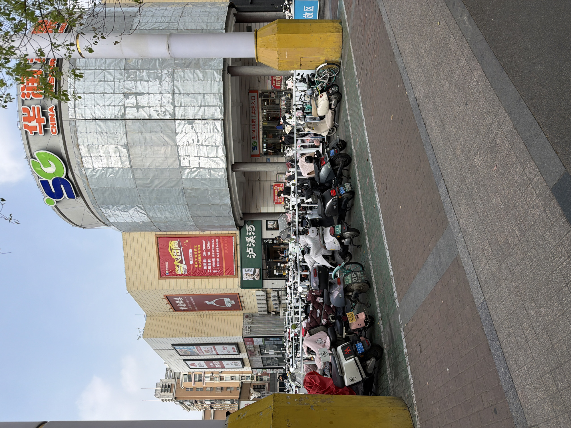 This image captures a vibrant urban scene in Jiangning District, China, on a clear, bright day. The central focus is a large commercial building, partially covered with reflective, square-patterned cladding, suggesting either construction, renovation, or a unique architectural design. A prominent "SG" logo in blue and green is visible on its rounded facade, along with the Chinese characters "华润苏果" (CR Suguo), identifying a supermarket chain, and the word "CHINA."
In front of the building, a long line of electric scooters, motorcycles, and bicycles are parked along a designated area on the sidewalk, marked by a sign reading "放区" (Parking Area). Several people are visible near the building's entrance, which is labeled "卖场入口" (Sales Floor Entrance), and among the parked vehicles. A large, yellow, concrete barrier or pillar stands prominently near the entrance.
To the right, a sequence of multi-story commercial buildings continues, adorned with various advertisements and signs. A large red banner advertises "大招商" (Big Investment Attraction) for "将军大道招商区" (Jiangjun Avenue Investment Area), detailing shop sizes (30-300m² for the first floor, 18-120m² for the second) and contact numbers for "顾经理" (Manager Gu) and "朱经理" (Manager Zhu). Another sign for "泸溪河" (Lu Xi He), possibly a brand or store, is also visible. The ground is paved with patterned gray and brown tiles for the sidewalk, transitioning to a darker road surface. Tree branches with green foliage appear in the upper left corner of the frame, hinting at nearby greenery. Visible vehicle license plates include numbers like "1672428" and "C103961".