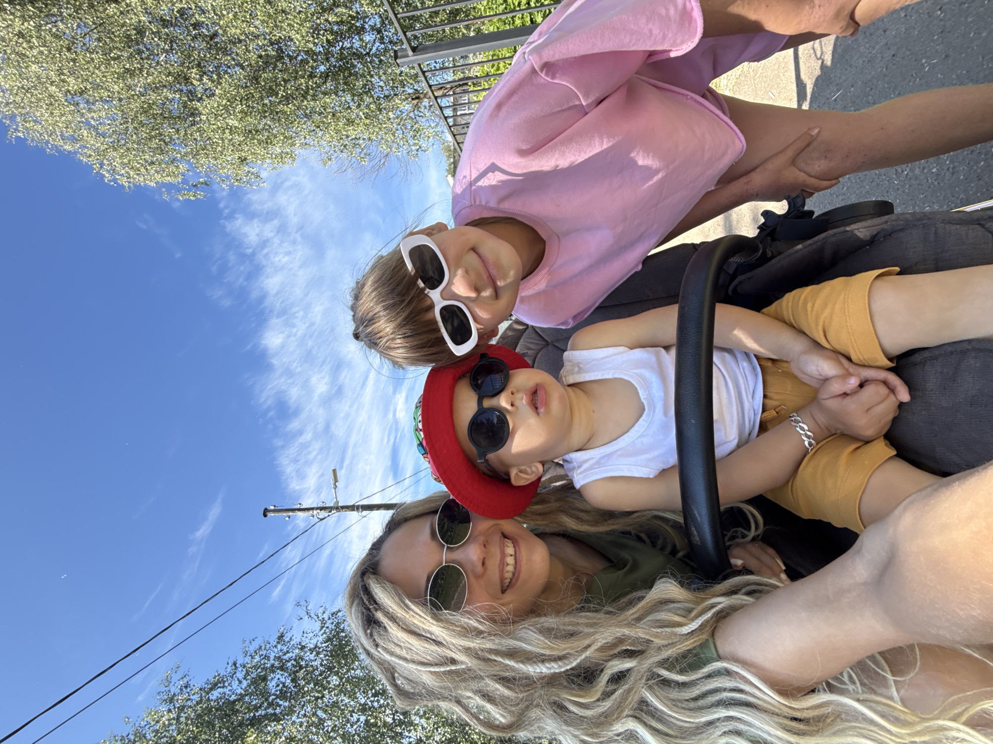 A mother and her two children are posing for a selfie outdoors on a sunny day. The mother, with long blonde dreadlocks and oval sunglasses, is on the left. To her right, a toddler wearing a red hat, round sunglasses, and a white tank top sits in a stroller. Next to the toddler, on the far right, is a young girl with brown hair and rectangular white sunglasses, wearing a pink sweatshirt. The background shows a blue sky with wispy clouds, trees, and a metal fence. The scene appears to be a street or park area in Rusanovo, Russia, with a power line visible overhead.
