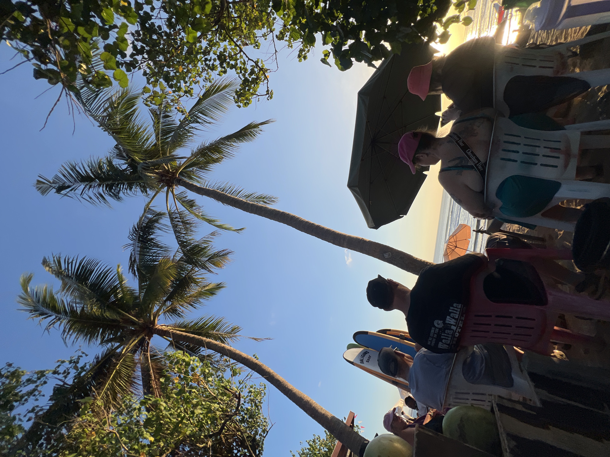 In Kuta, Indonesia, on a beach, several people are seated at tables under palm trees, enjoying the sunset. The image captures a relaxed atmosphere, with the ocean and sky as a backdrop. Two individuals are seen in the foreground, facing away from the camera, while others are seated around them. Surfboards are visible, suggesting the proximity to a surfing spot. The lighting indicates it's either sunrise or sunset, casting a warm glow on the scene. The environment is tropical, with lush vegetation and the sandy beach characteristic of the location.