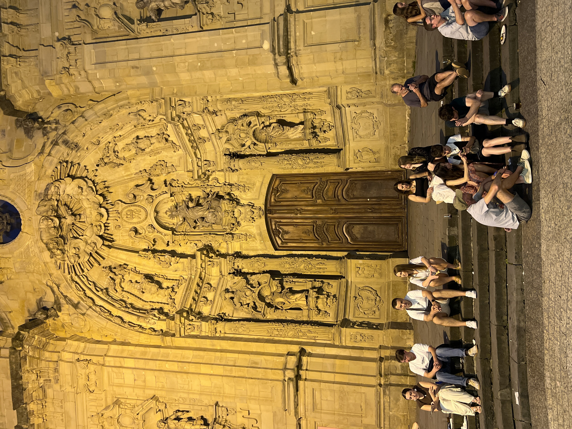 The image shows the ornate facade of the Church of San Sebastián Mártir in San Sebastián, Spain, at night. The church's facade is illuminated, highlighting its intricate stone carvings and sculptures. In front of the church, on a set of wide stone steps, several groups of people are seated, resting and socializing. Some individuals are looking at their phones, while others are engaged in conversation. The atmosphere appears relaxed, with people enjoying the evening outside the historic building. The steps are made of dark stone, and the ground in front of them is paved. The lighting casts a warm yellow glow on the scene.