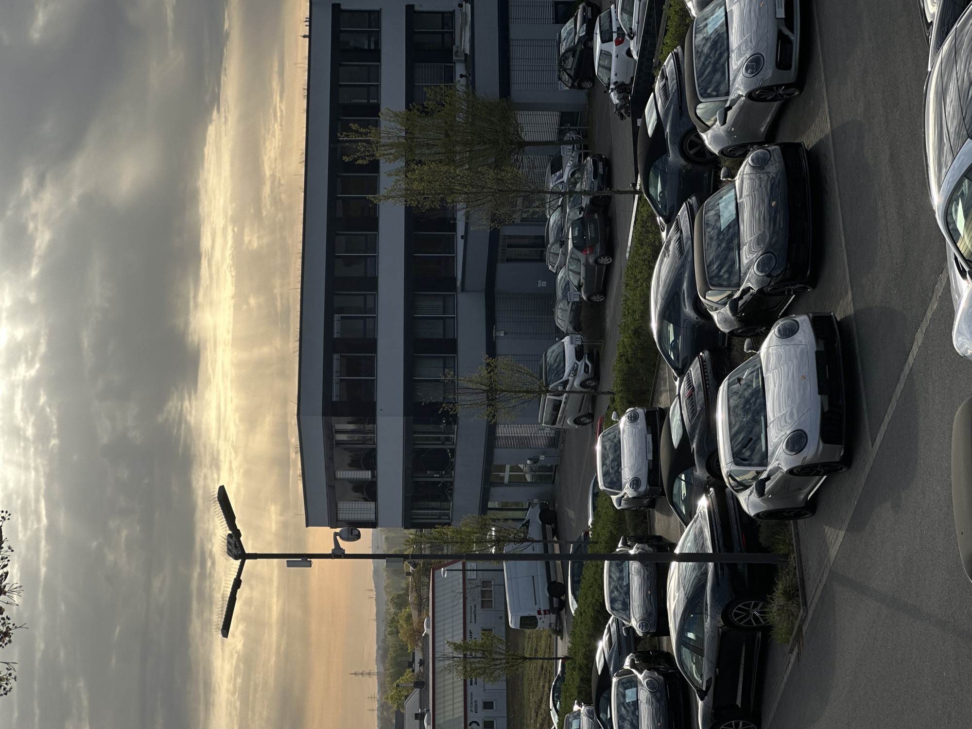 The image shows a parking lot in Holzwickede, Germany. The parking lot is adjacent to a modern building with multiple floors and windows. The sky is cloudy with a yellow glow from the sun near the horizon. There are rows of cars parked neatly in the lot, predominantly Porsche models. A lamppost with a camera is visible on the left side of the image.