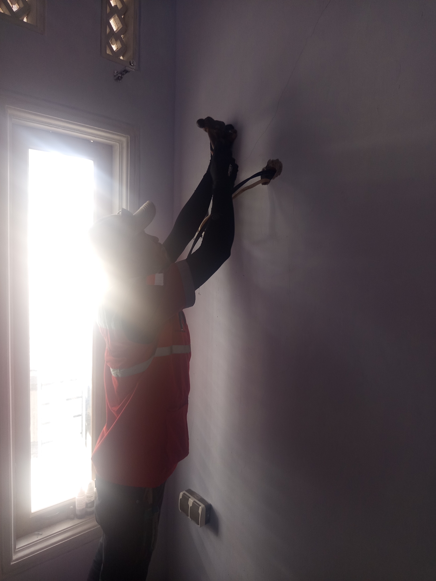 An Indonesian electrician, wearing an orange and reflective vest and dark pants, is working on electrical wiring near a window in a room. The electrician is facing the wall, with their back partially to the window. Two electrical wires are visible extending from the wall, and the electrician is holding them, appearing to be in the process of connecting them. A window with white blinds is to the left of the frame, letting in bright sunlight that illuminates the room and casts a shadow of the electrician on the wall. To the right, a wall-mounted electrical outlet is visible. The room appears to be in a residential or commercial building in Sukoreno, Indonesia.