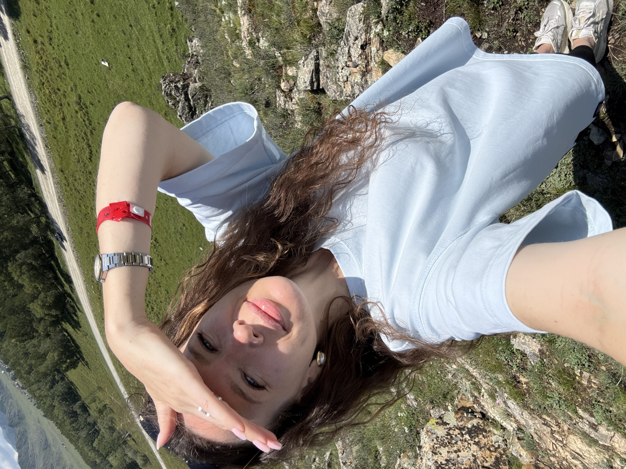 A young woman lies on her side, holding her hand to her forehead. She wears a light blue t-shirt and white sneakers. A red wristband is visible on her left arm, along with a silver watch. She has long, brown, wavy hair and is looking towards the camera with a slight smile. The setting appears to be a grassy, rocky hillside with a dirt road winding through it in the background. The sun is shining, suggesting it is daytime. The overall mood seems relaxed and happy.