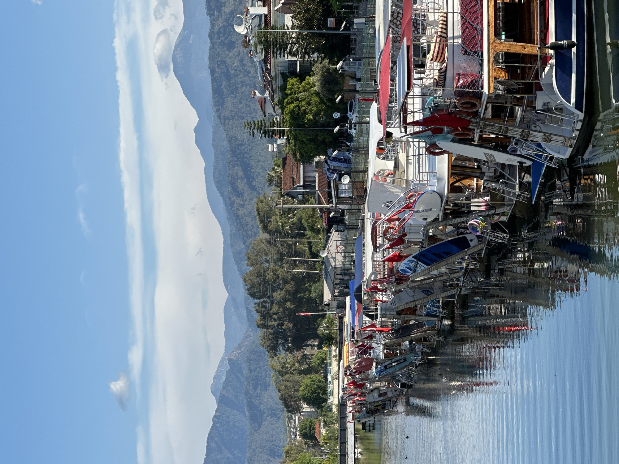 The image depicts a waterfront scene in Fethiye, Turkey. A row of boats is docked along the harbor, their reflections visible in the water. Mountains rise in the background, and buildings line the shoreline. The sky is blue with scattered clouds.