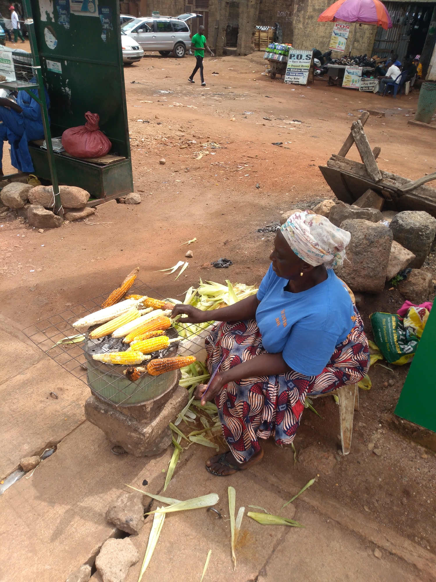 This is an outdoor scene in Jos, Nigeria, featuring a woman preparing corn. The woman, wearing a blue t-shirt and a colorful patterned skirt with a headscarf, is seated next to a charcoal grill where several ears of corn are being roasted. The corn is arranged on a metal grate over the grill, which is placed on top of stacked stones. The ground is covered in reddish-brown earth and concrete. In the background, various structures and objects suggest a market or street vendor setting, including a green metal kiosk, a silver SUV, and a street cart with drinks and signage. The overall environment appears to be an urban, outdoor marketplace.