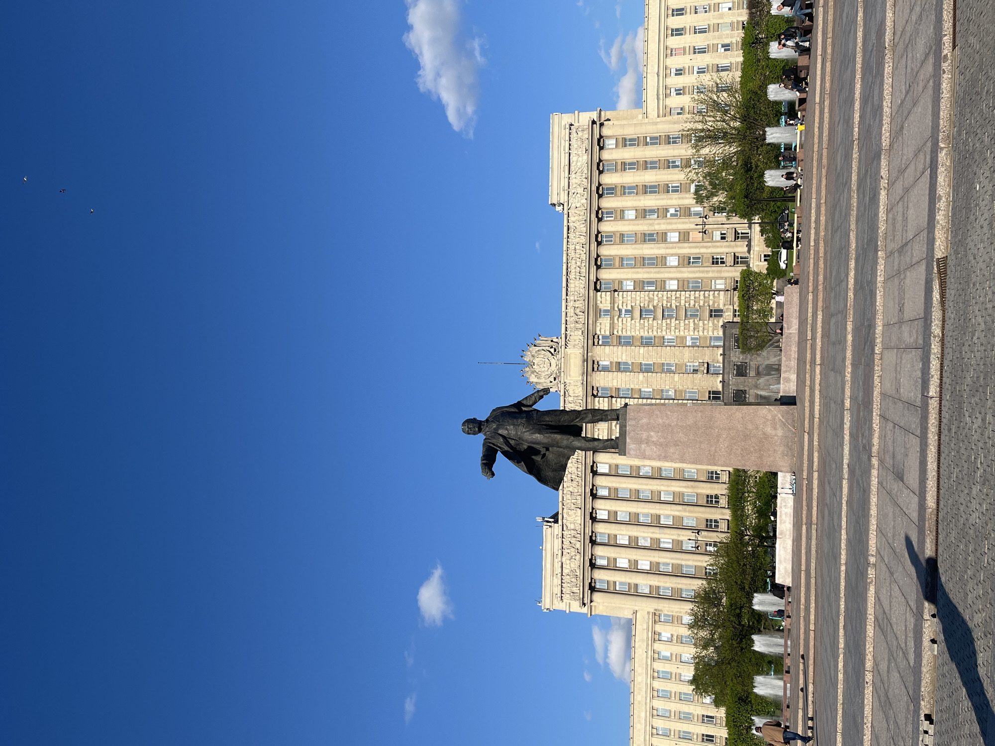 The image shows a statue of Vladimir Lenin in Saint Petersburg, Russia. The statue stands in front of the House of Soviets building. The sky is clear and blue with a few clouds. The image is taken at ground level. There are multiple buildings and trees. The lighting suggests it is daytime.