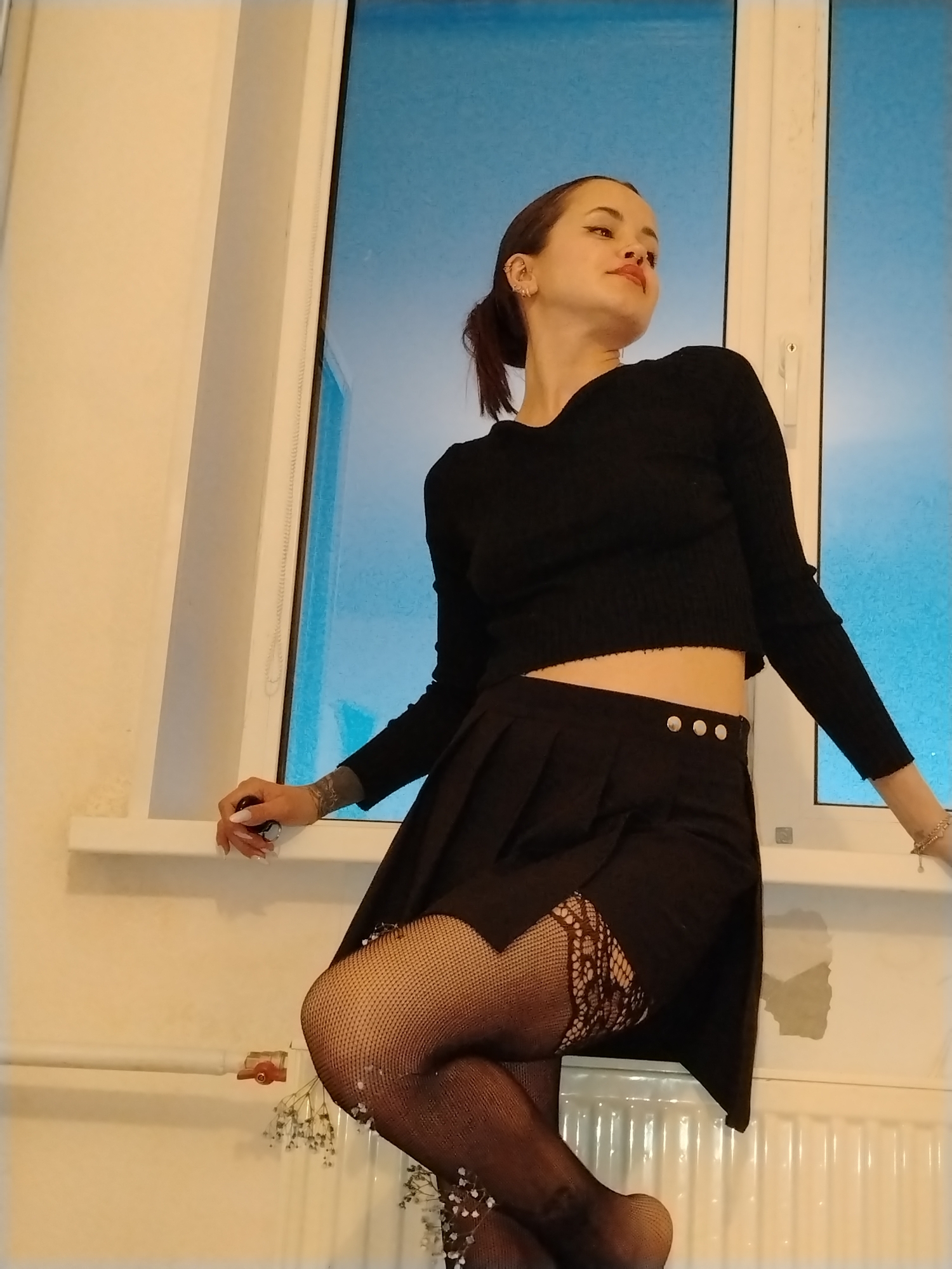 A woman is posing in front of a window. She has dark hair pulled back in a ponytail, dark eye makeup, and several ear piercings. She is wearing a black long-sleeved cropped sweater, a short black skirt with button details at the waist, and black fishnet stockings with lace detail at the thigh.
The background is a white-framed window with a blue-grey hue visible outside. The woman is resting her hands on the window ledge. A white radiator is visible beneath the window, with a small bunch of baby's breath flowers tucked beside the radiator valve.
No specific location or city in Russia is discernible from the image.