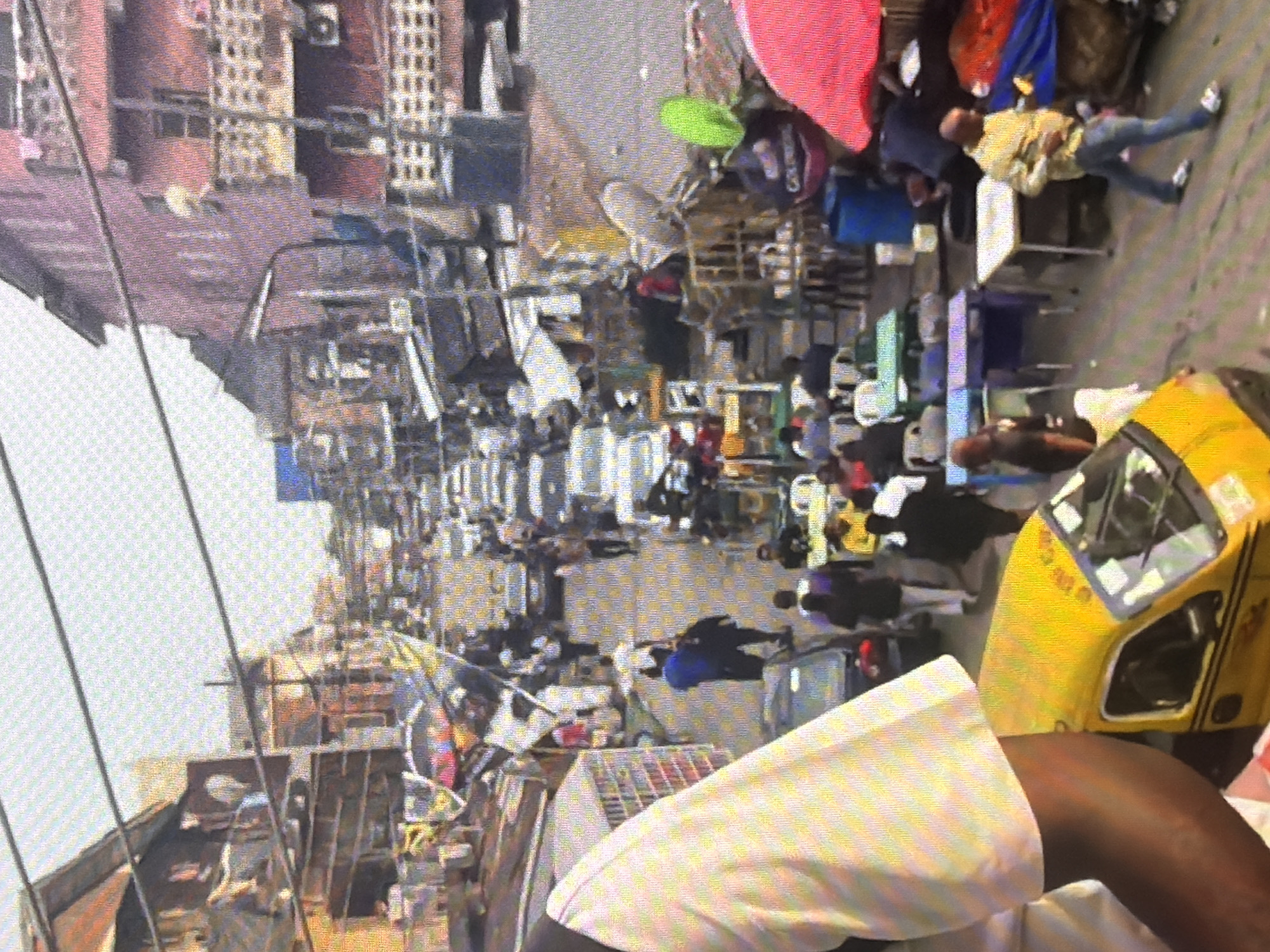 The image depicts a bustling street scene in Bukuru, Nigeria, likely a market area. The street is narrow and filled with people and vehicles, suggesting a busy commercial environment. Buildings line both sides of the street, some appearing to be multi-story structures with balconies. The overall atmosphere is one of a lively, crowded urban setting.
In the foreground, a yellow auto-rickshaw, also known as a "keke," is visible, with its front section taking up a significant portion of the left side of the frame. A person's arm, appearing to be dark-skinned and wearing a white, striped garment, is also prominent in the lower left corner.
Further down the street, a variety of people are engaged in different activities. Some individuals appear to be walking, while others are gathered around stalls or tables. There are also several cars and motorcycles parked or moving along the street. The presence of numerous people suggests a public gathering or market activity.
The environment appears to be somewhat cluttered, with various objects and structures visible throughout the scene. These could include market stalls, makeshift shelters, and assorted items for sale. The sky is visible in the upper portion of the image, appearing overcast or hazy. The scene is densely populated, indicating a high level of human activity and a typical representation of a busy commercial street in Nigeria.