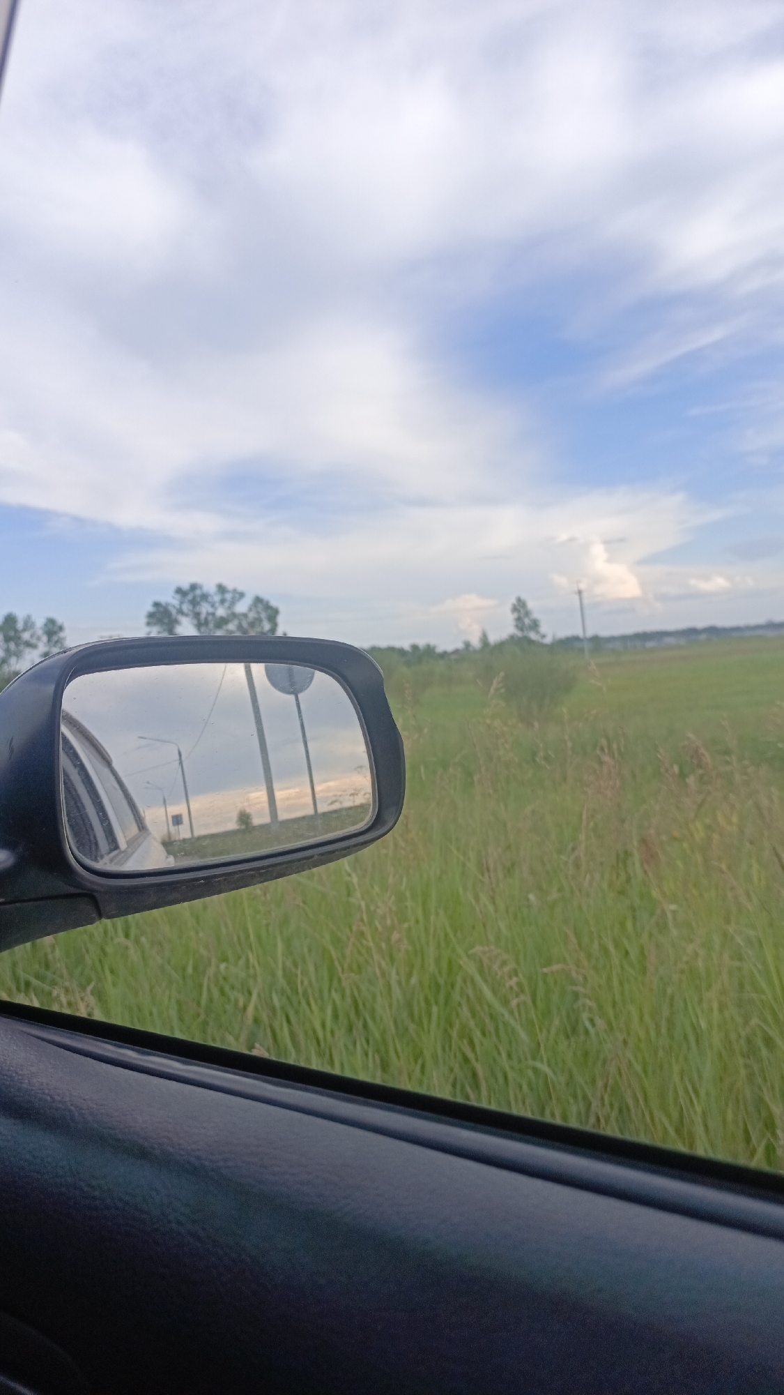 The image shows a view from inside a car looking out the side window and into the side-view mirror. The car is on a road next to a field of tall green grass. In the mirror, a road with two streetlights and a round traffic sign is reflected. The sky is blue with white clouds. The scene appears to be in a rural or suburban area, possibly on the outskirts of Angarsk, Russia, given the presence of typical Russian streetlights and road signs. There are no people visible in the image.