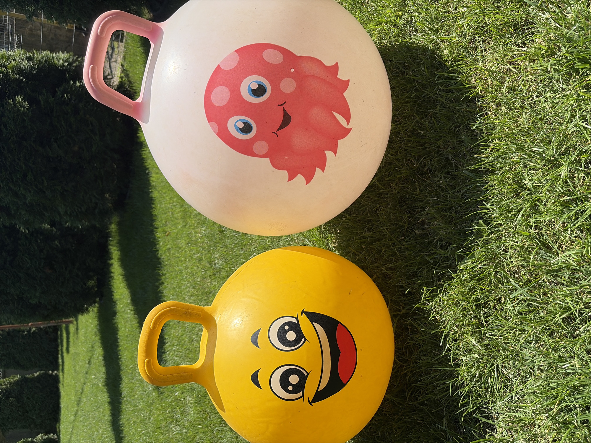 Two inflatable hopper balls, one yellow with a smiley face and a pink octopus on a white ball, sit on a grassy lawn. The balls are positioned side-by-side, with the yellow ball on the left and the white ball on the right. The lawn appears to be well-maintained and is partially shaded by unseen trees. In the background, dark green bushes or trees form a dense backdrop. The scene is lit by bright sunlight, casting shadows on the grass. The image was taken in Oxentea, Moldova.